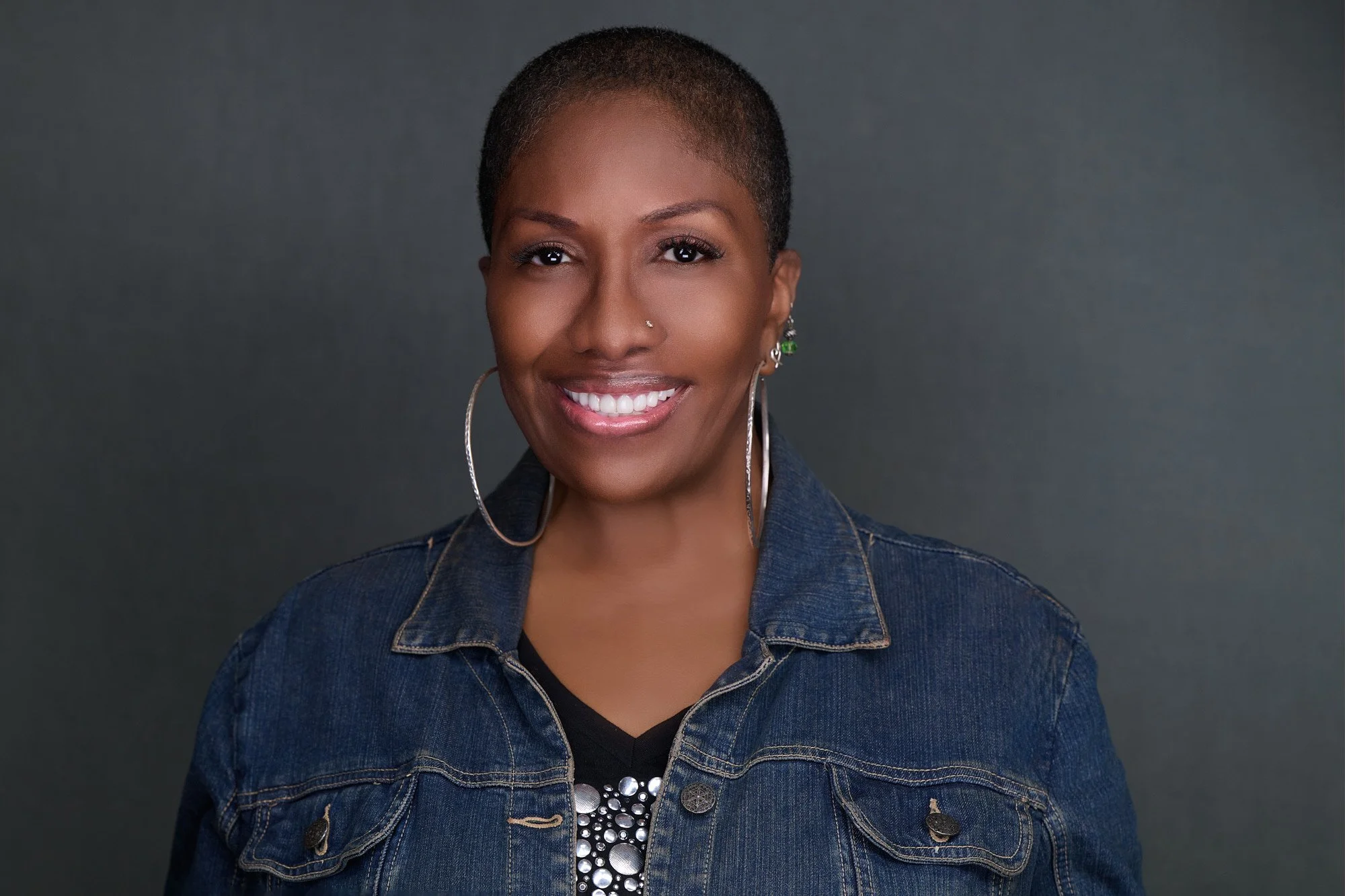 Corporate headshot of a woman in a denim jacket and hoop earrings on a charcoal backdrop, Orange County team headshot session