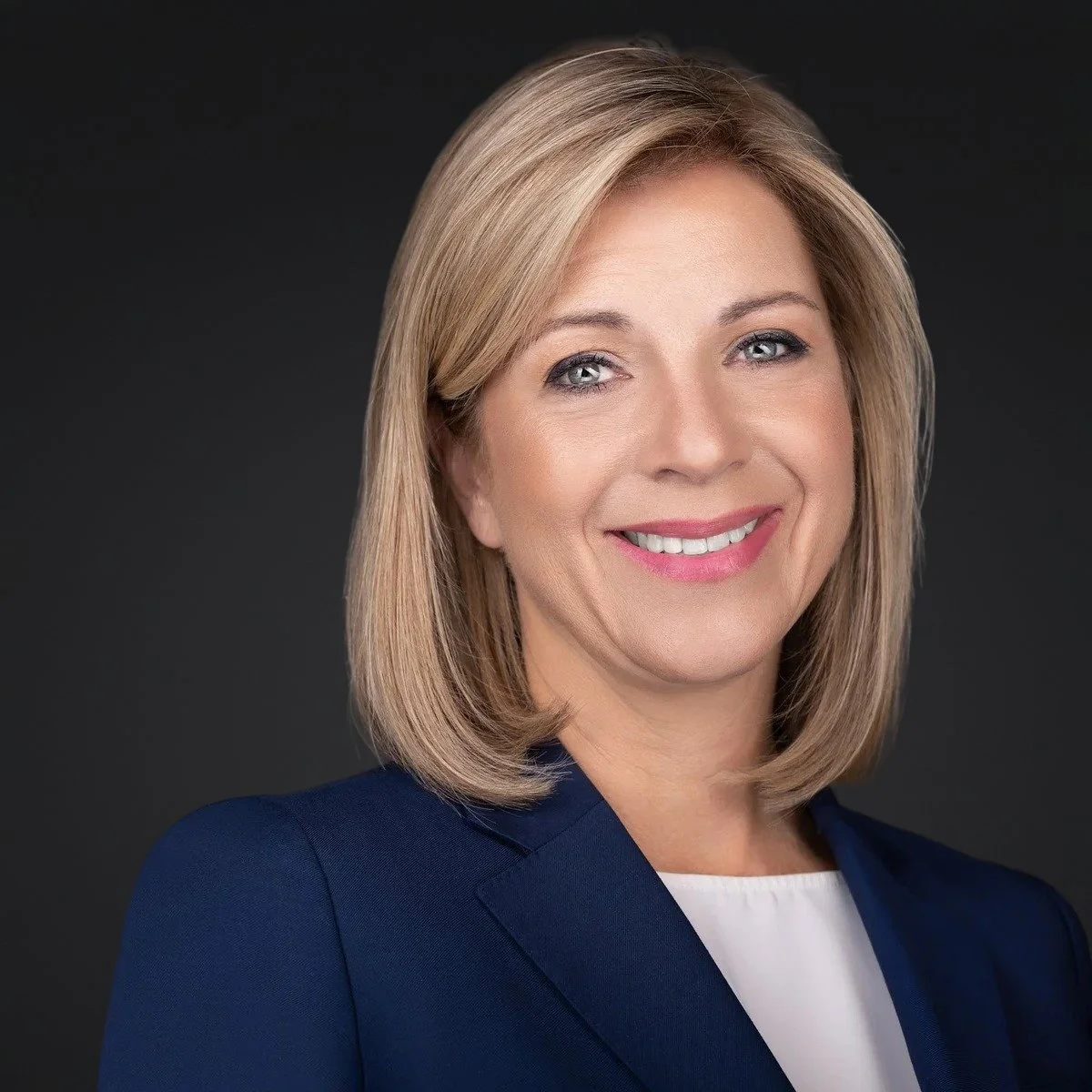 Business headshot of a smiling woman with shoulder-length blonde hair in a navy blazer and white top, on a dark gray background.