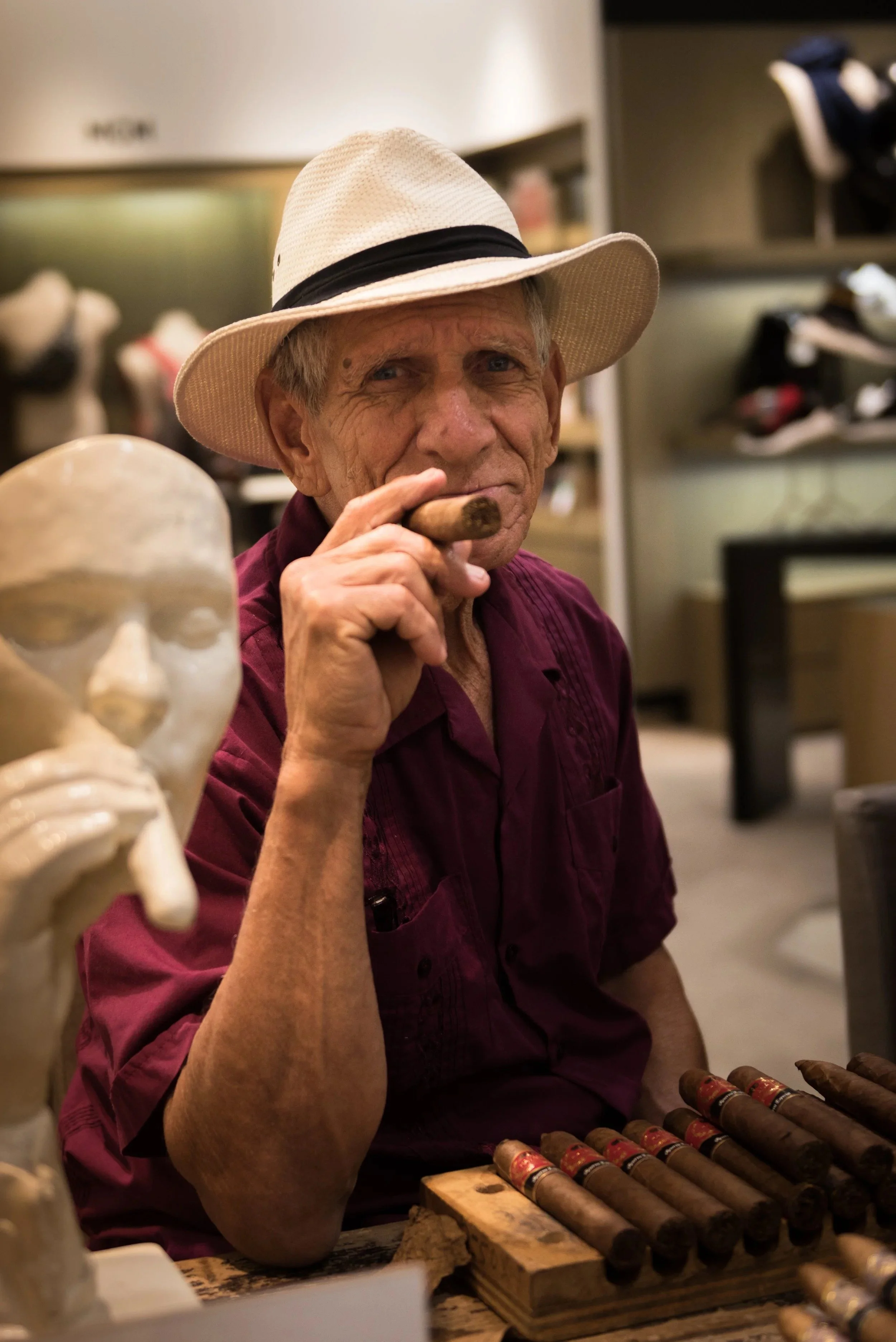 Environmental portrait of an older man in a straw hat smoking a cigar at his shop.