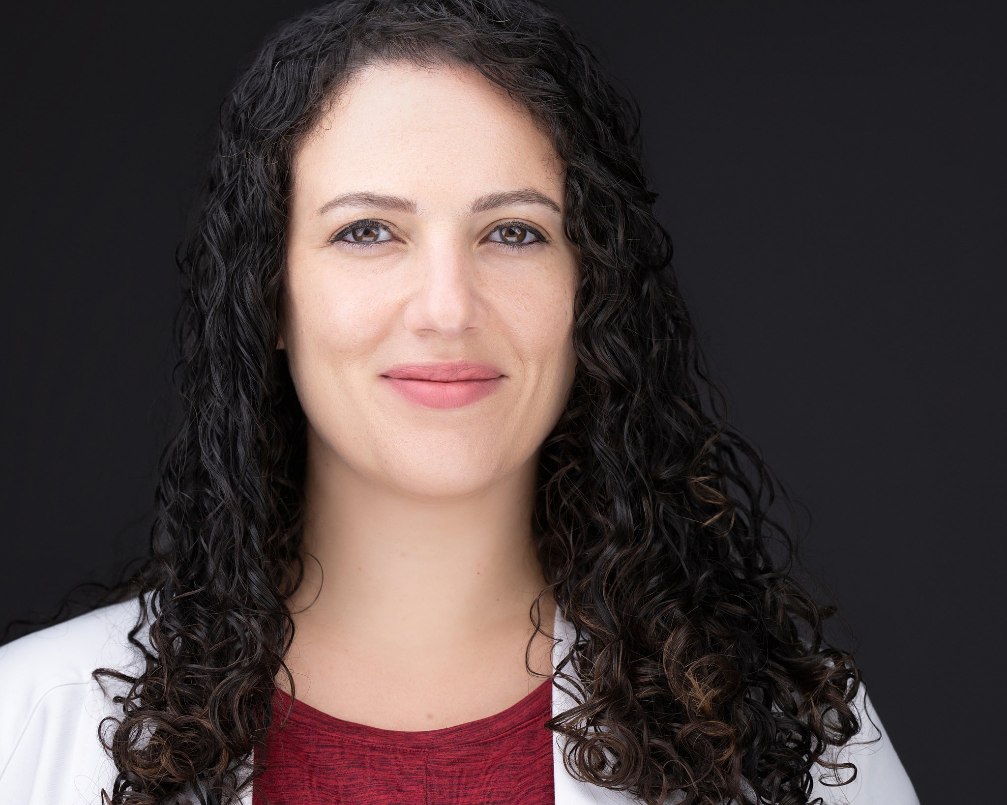 Medical headshot of a woman with long curly dark hair in a white coat over a burgundy top, photographed on a dark background.