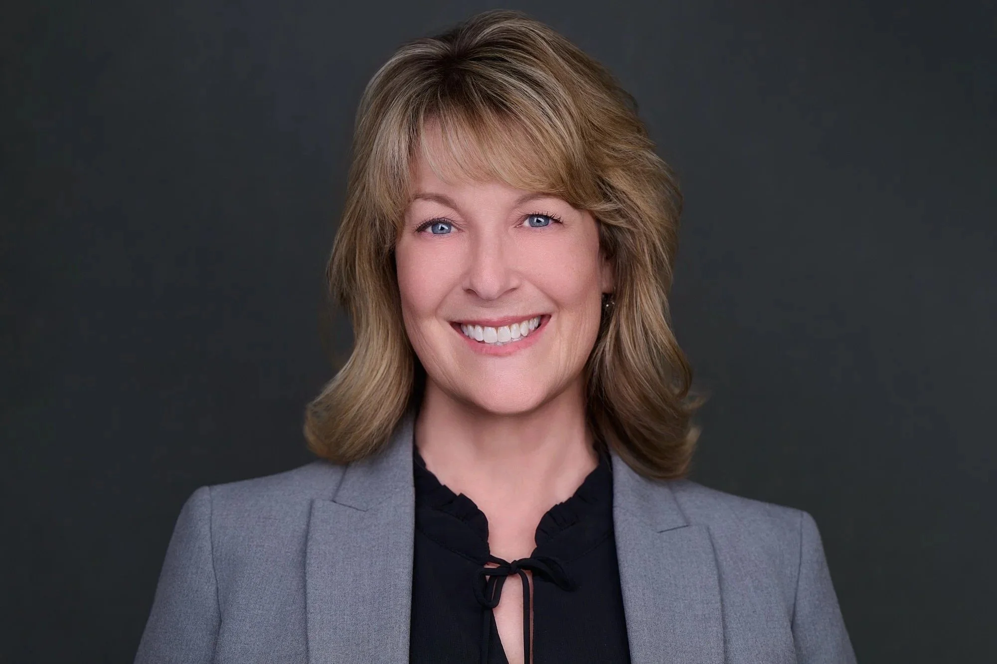 Corporate headshot of a smiling woman in a gray blazer on a charcoal background.