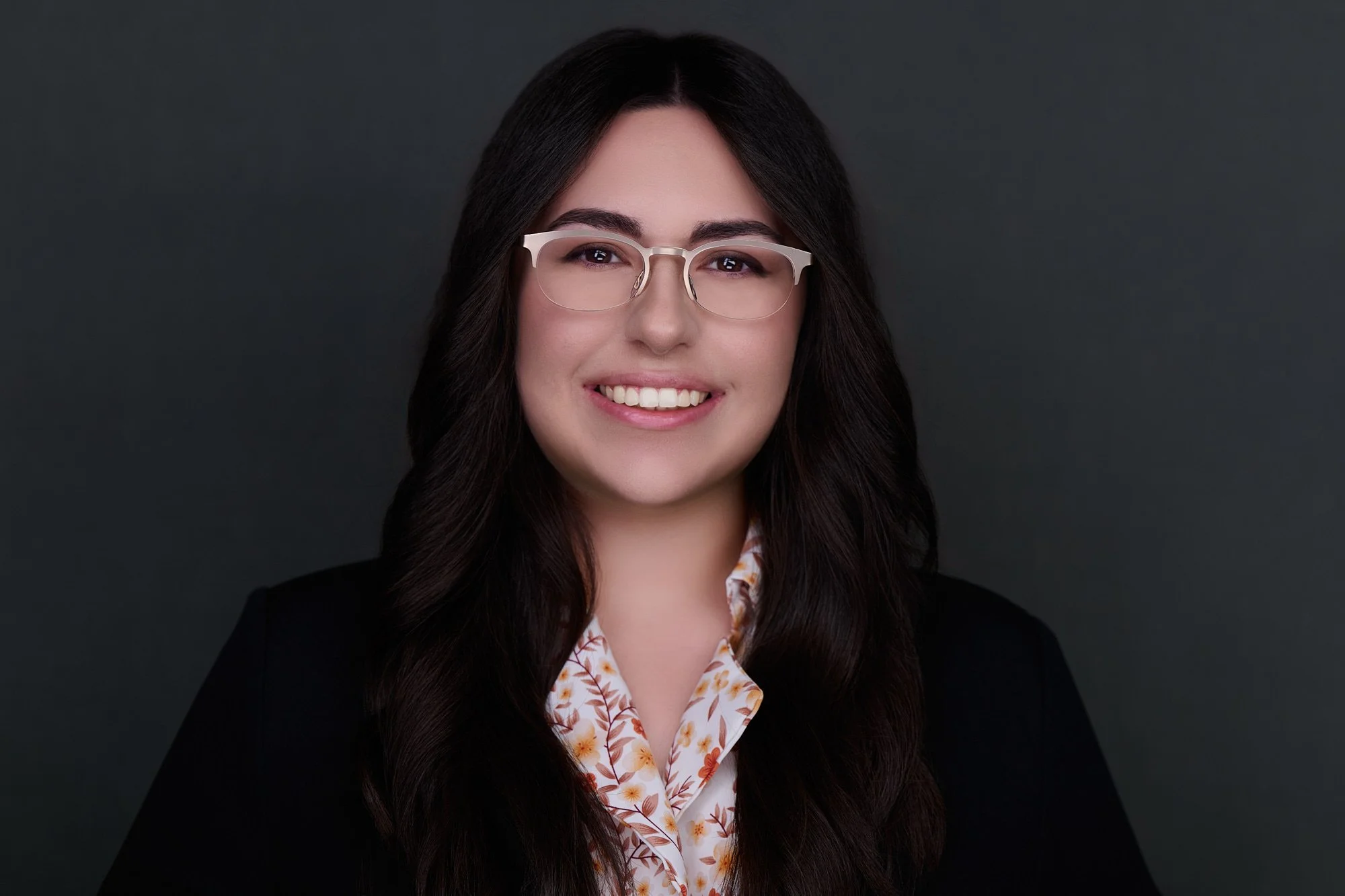 Corporate headshot of a woman with glasses in a floral blouse and black blazer on a charcoal backdrop, Orange County team headshot session