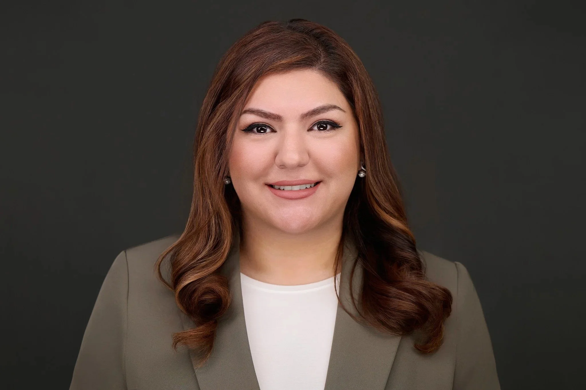Real estate headshot of a smiling woman with long auburn hair in a taupe blazer on a dark gray background.
