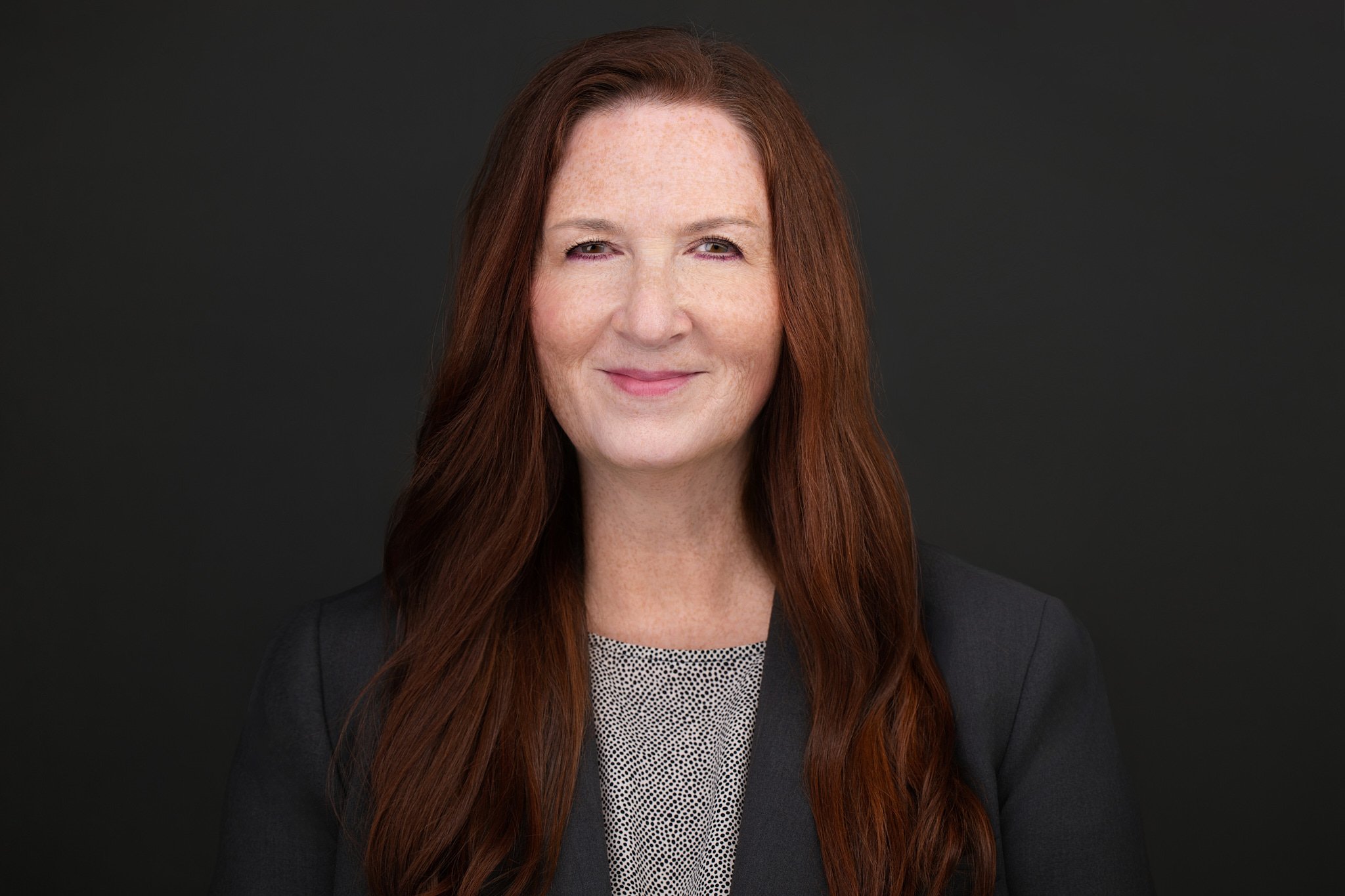 Business headshot of a woman with long auburn hair in a charcoal blazer, photographed on a dark gray background.