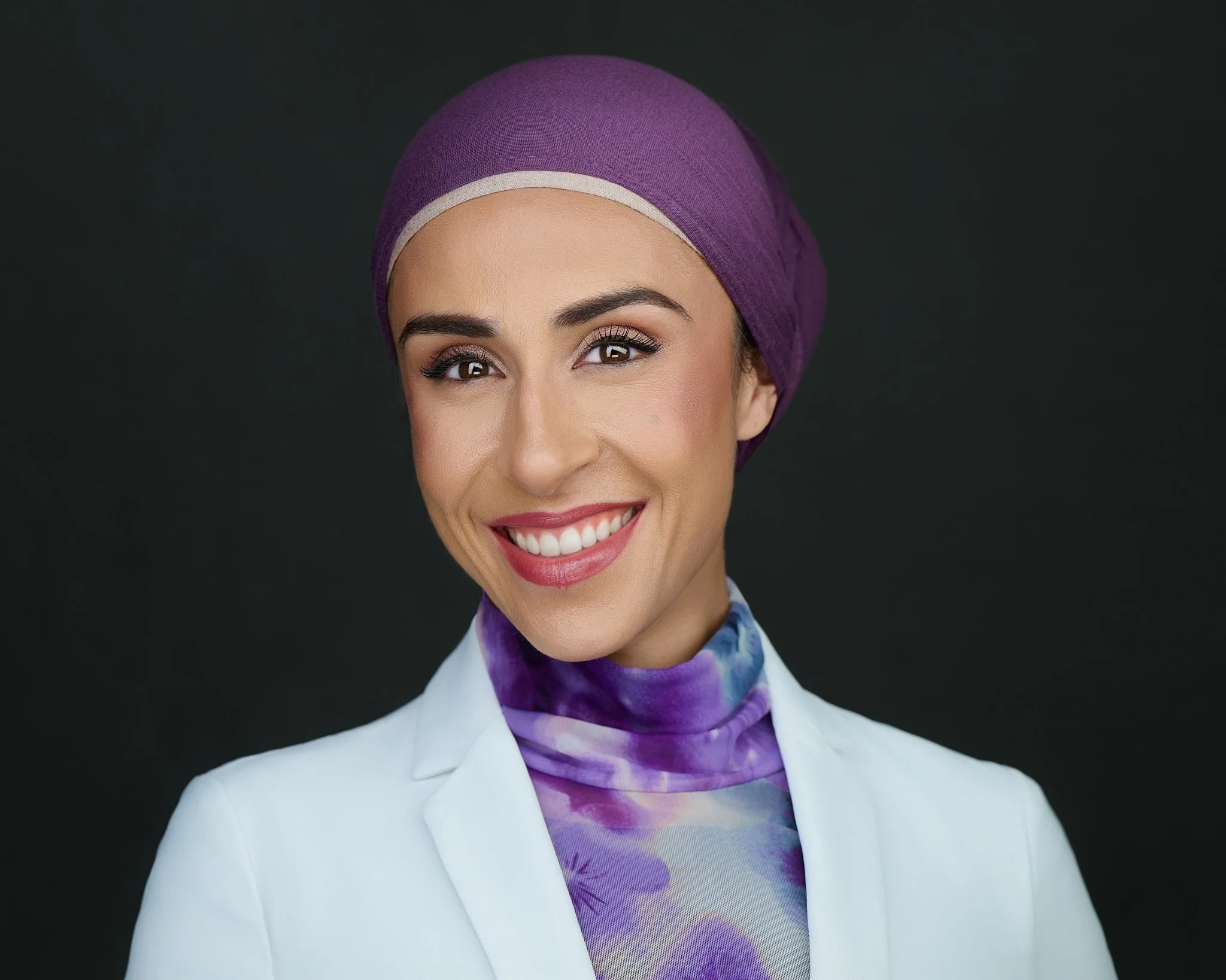 Studio headshot of a woman in a purple head wrap and white blazer against a black background.
