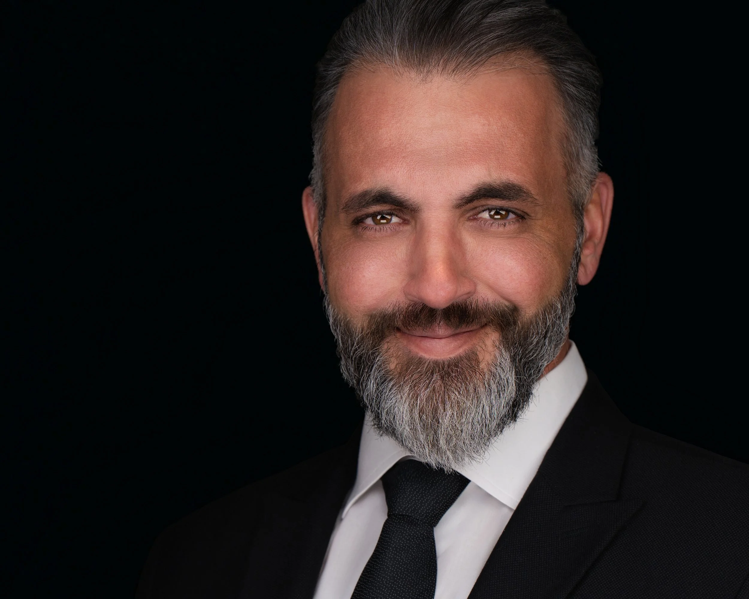 Executive headshot of a man with a grey beard in a black suit, white shirt and black tie on a black background.