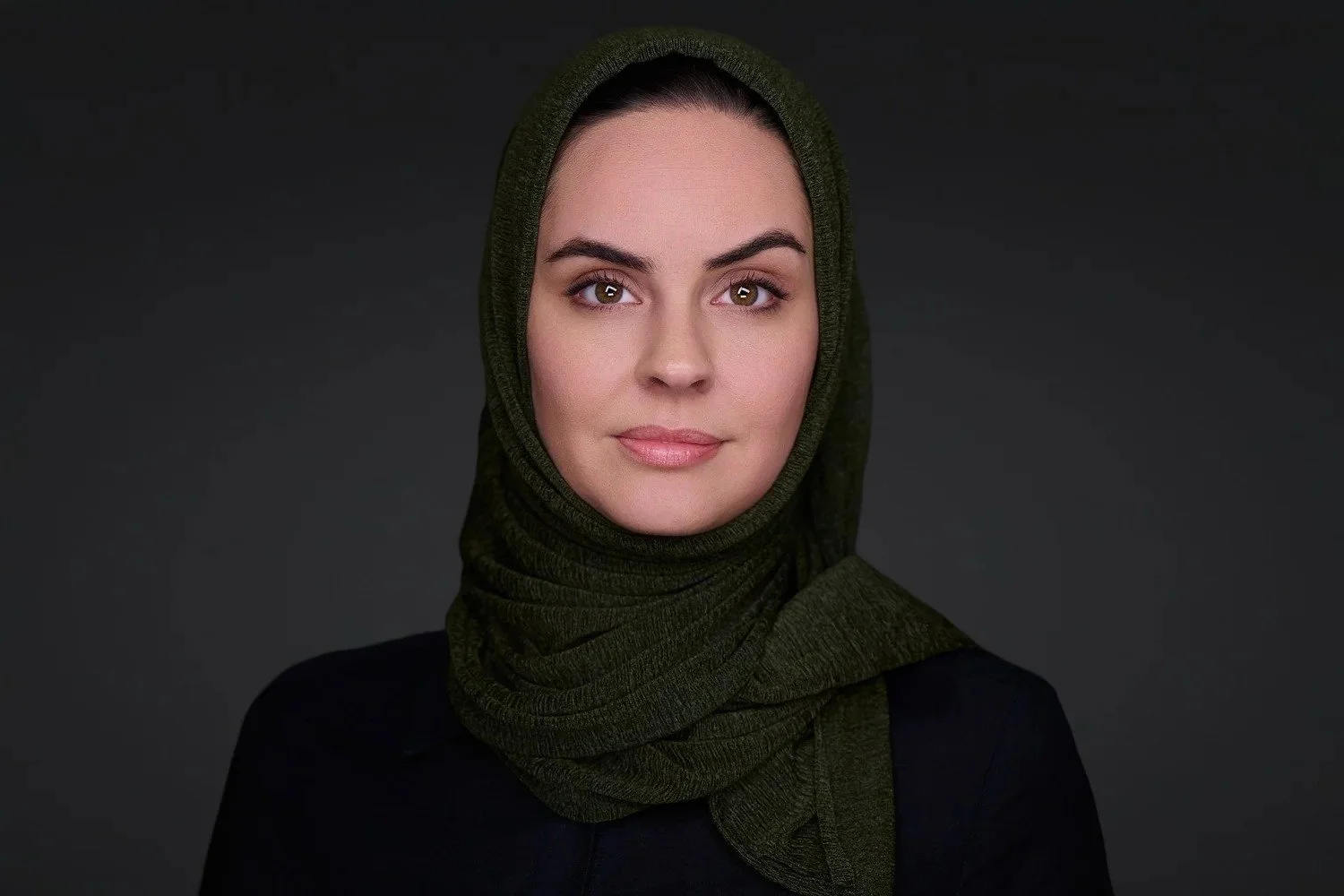 Professional headshot of a woman wearing a hijab and a dark blazer on a charcoal background