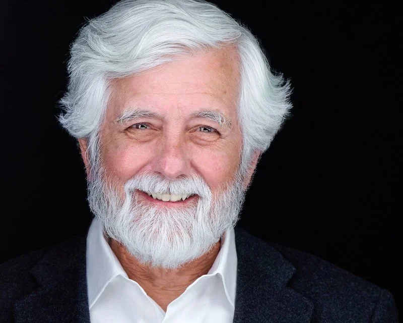 Professional headshot of a man with white hair and a full white beard in a navy jacket and white shirt, photographed on a black background.
