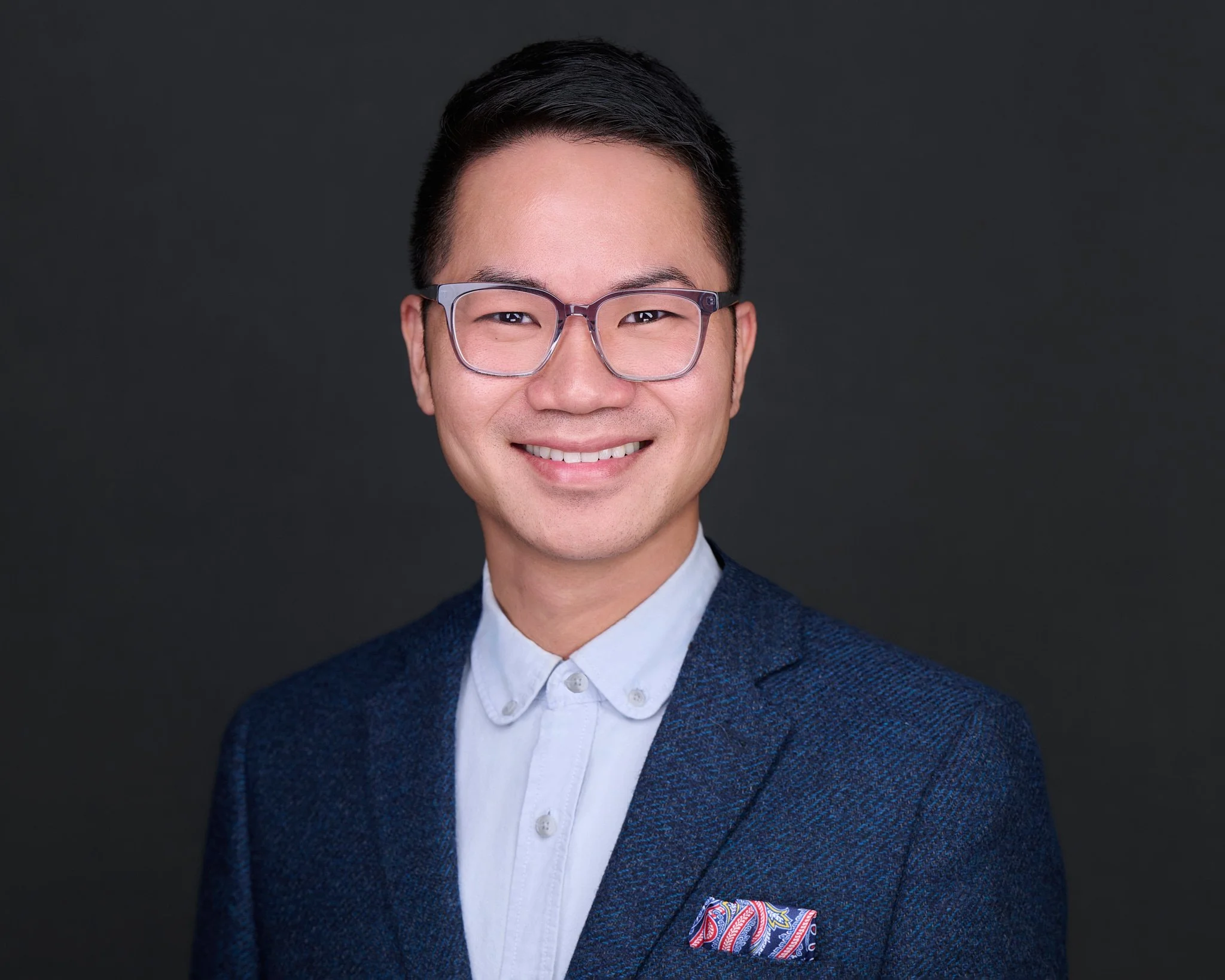 Studio headshot of a man in a navy tweed jacket and glasses, with a paisley pocket square, against a dark grey background.