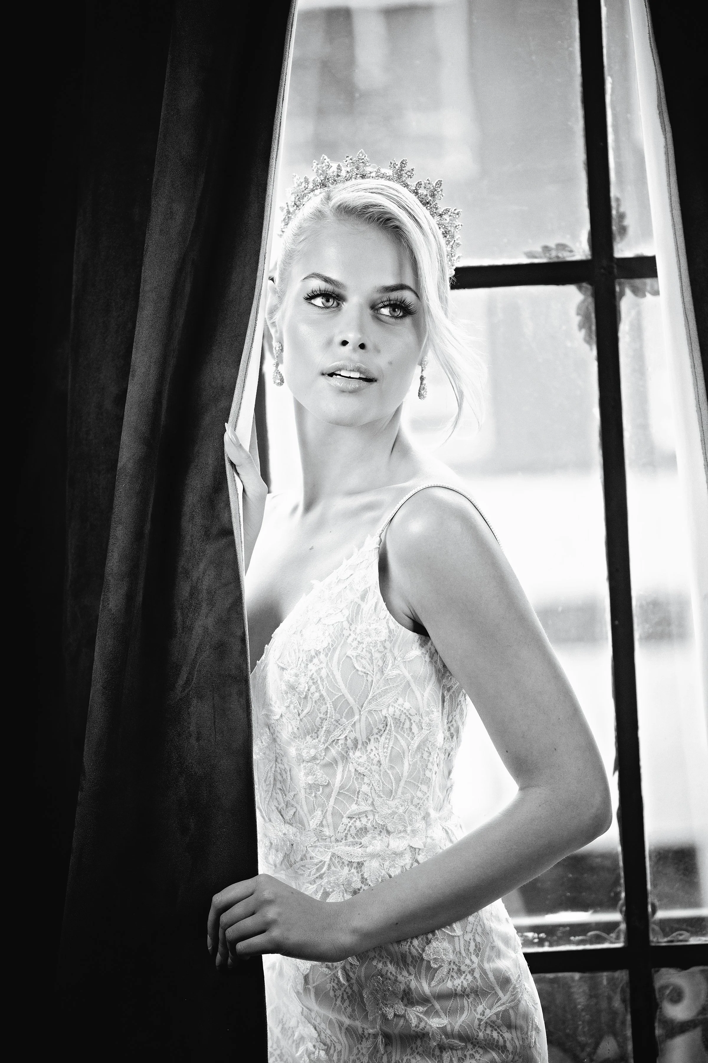 Black-and-white portrait of a woman in a lace gown and tiara by a window