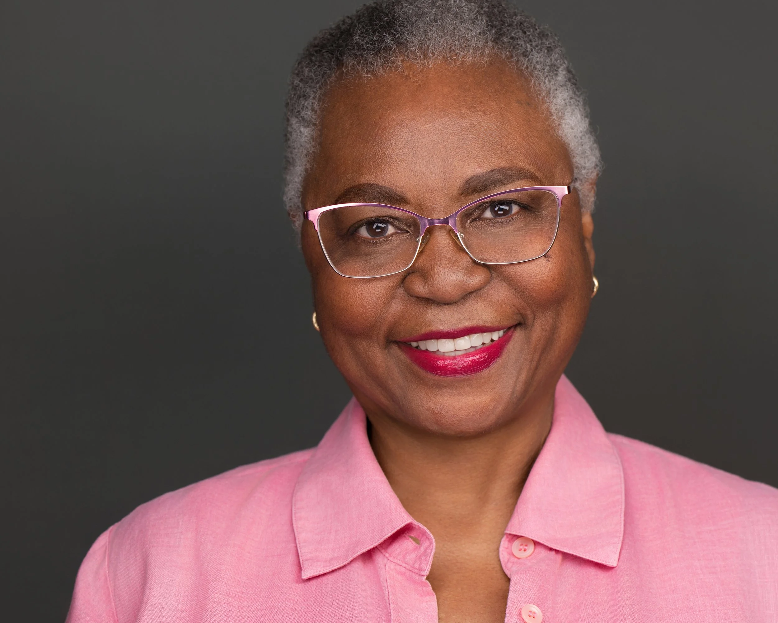 Studio headshot of a woman with short grey hair and pink glasses, wearing a pink linen shirt against a grey background.