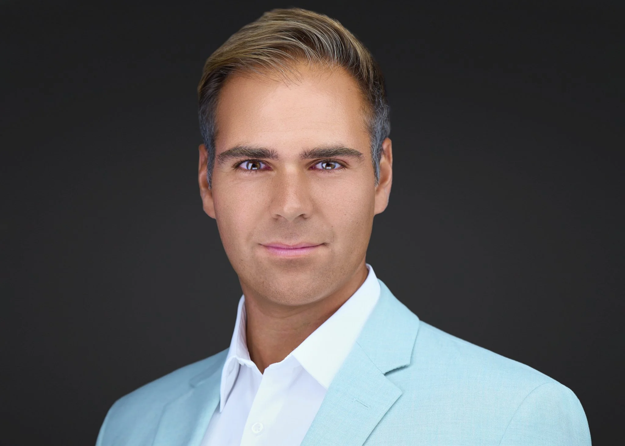 Executive headshot of a man in a light blue blazer and white open-collar shirt, photographed on a dark gray background.