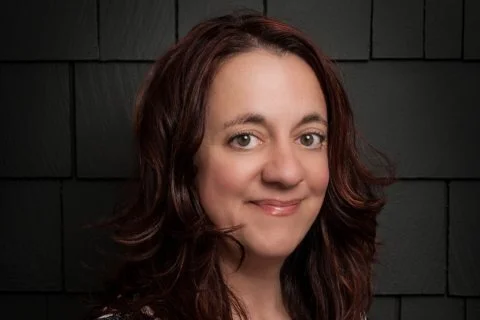 Portrait of a person with long brown hair, smiling slightly, against a dark tiled background.