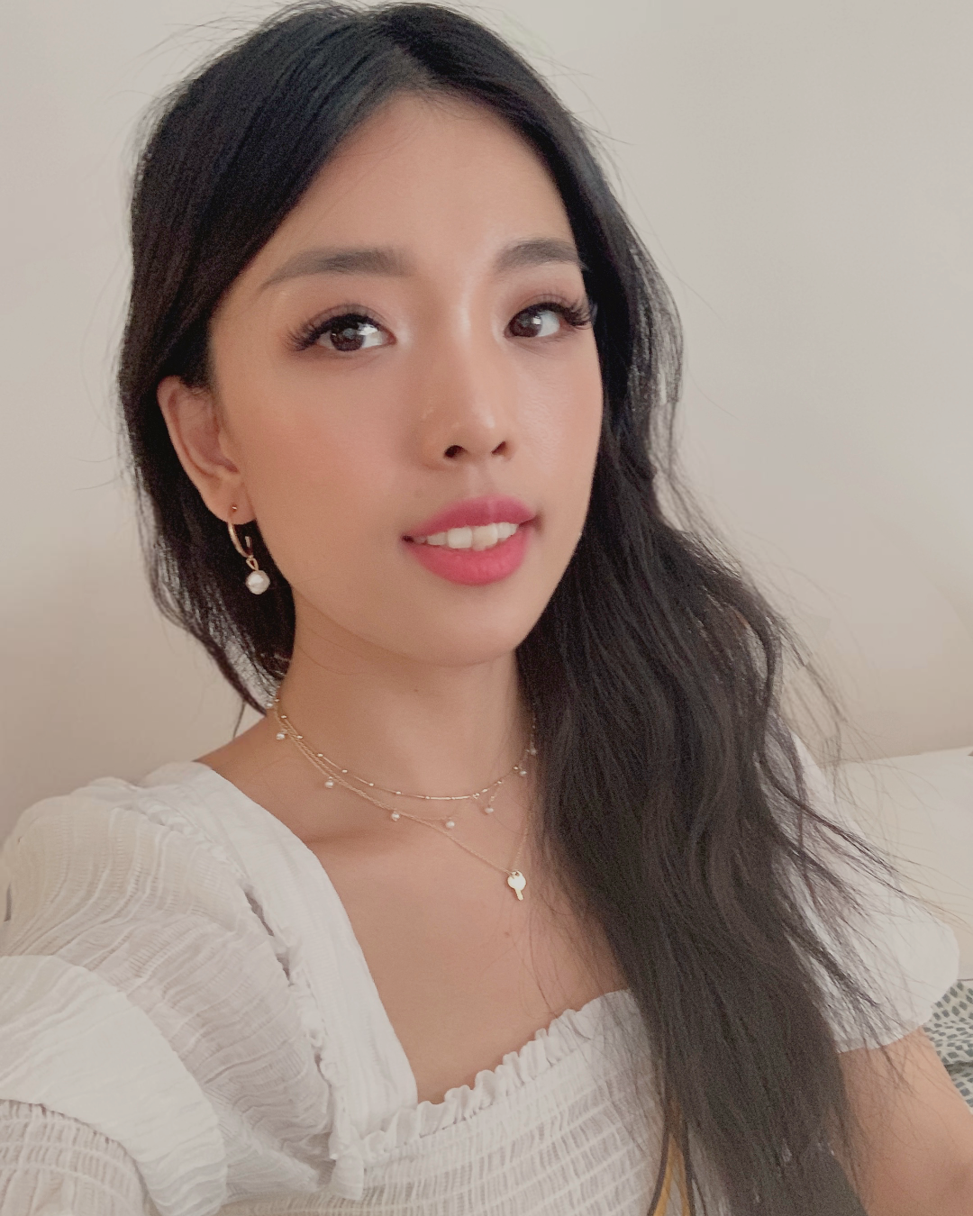 A young woman with long black hair, wearing makeup, earrings, and layered necklaces, taking a selfie indoors against a plain background.