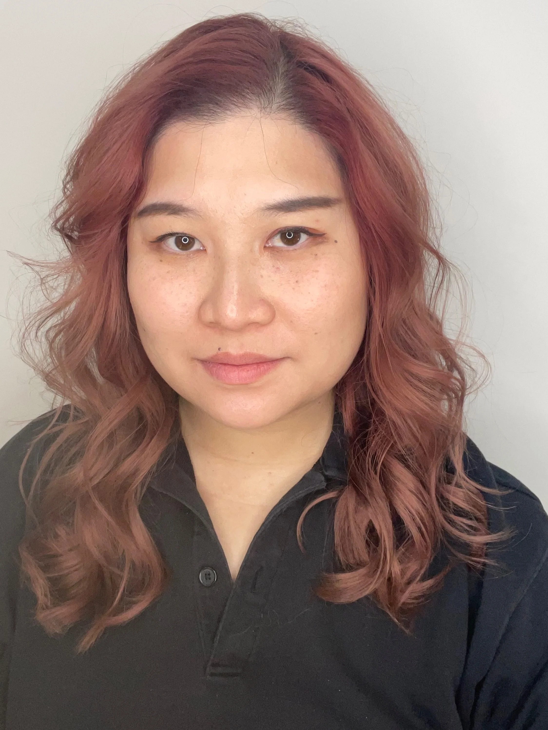 A woman with shoulder-length, wavy reddish-brown hair wearing a black collared shirt, looking at the camera against a plain light background.