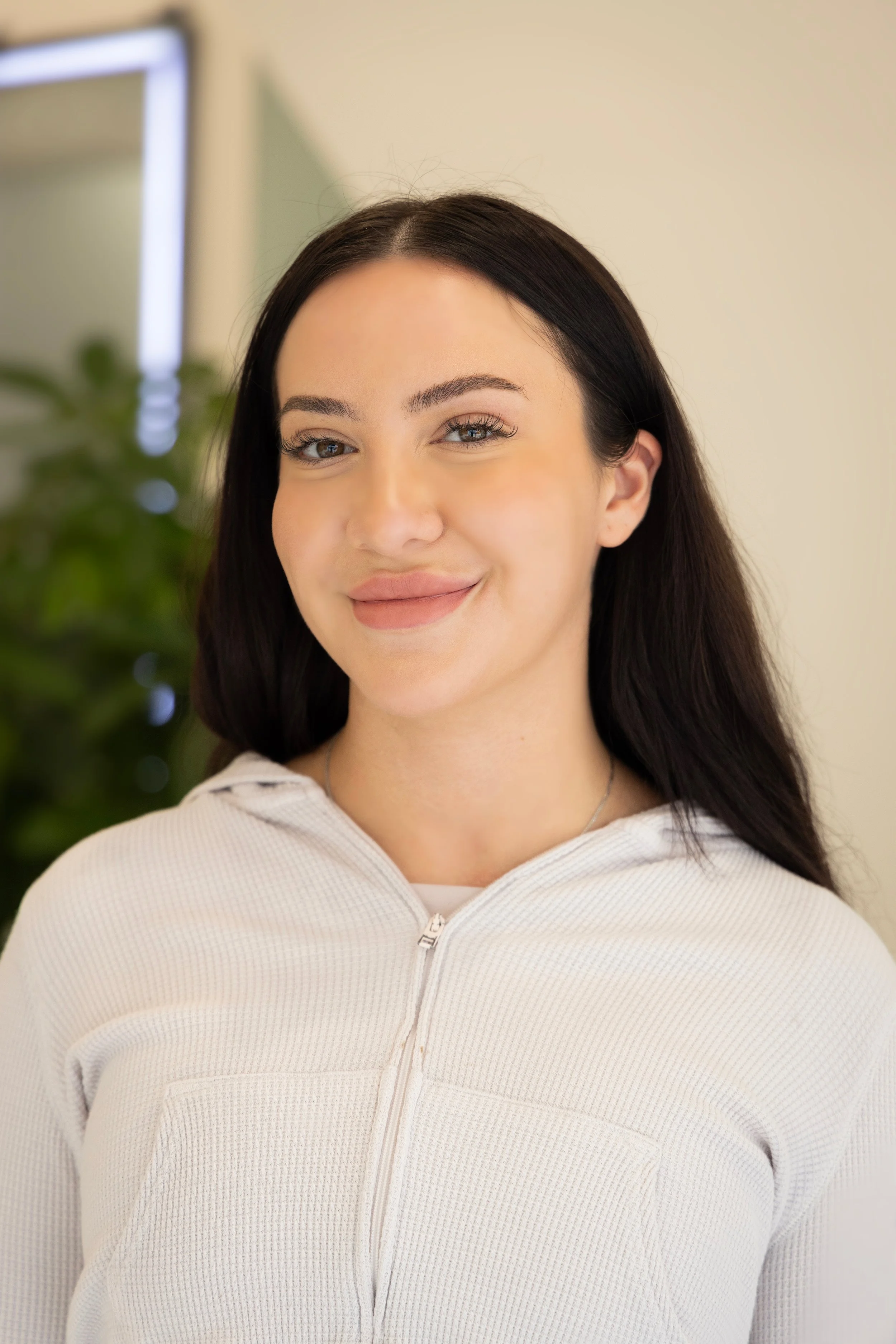 A woman with long dark hair, light skin, and a friendly smile, wearing a white zip-up top, standing indoors with a blurred background and a plant.