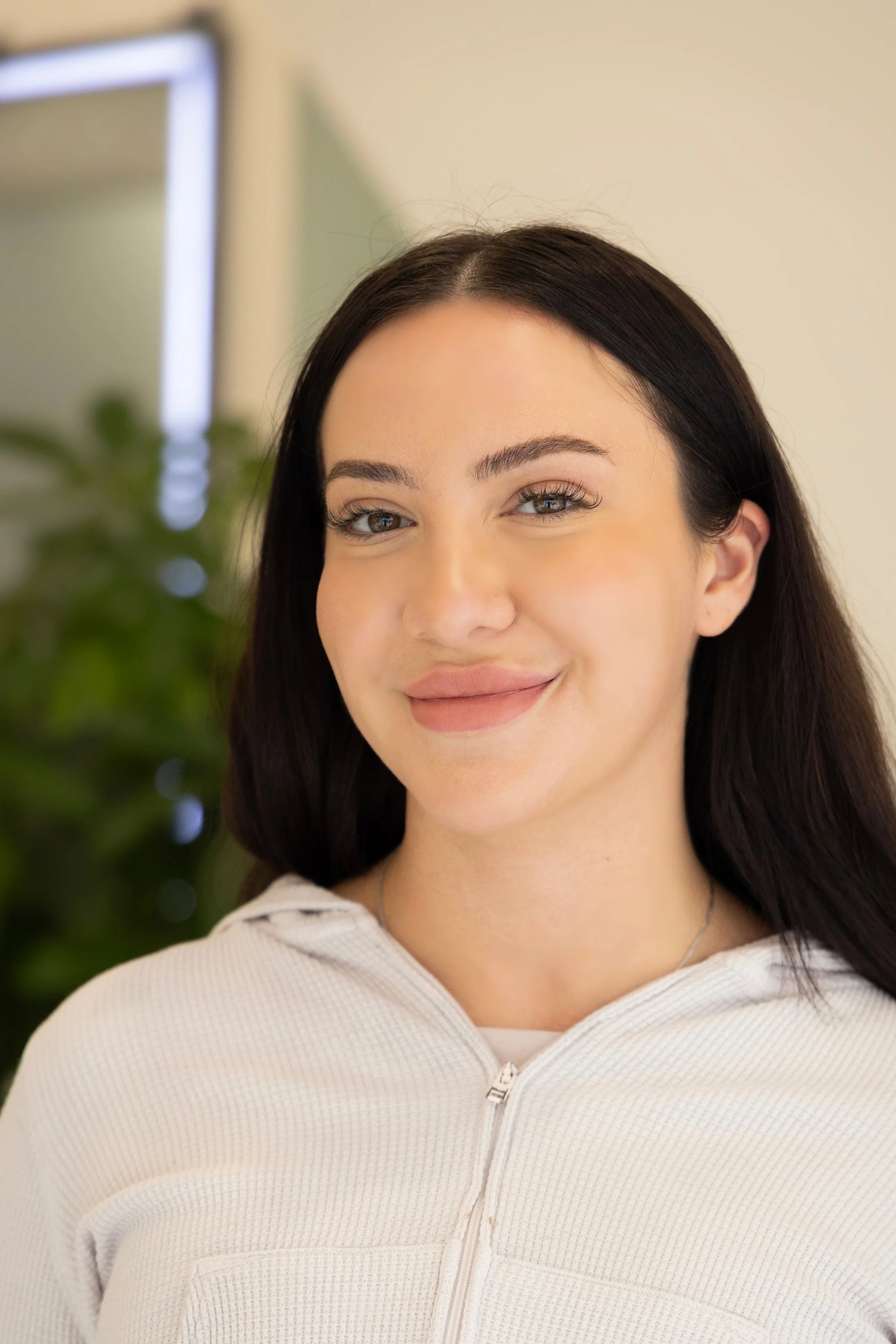 Close-up of a woman with long dark hair and light skin, smiling, wearing a white zip-up top, with a blurred background including a green plant and a beige wall.