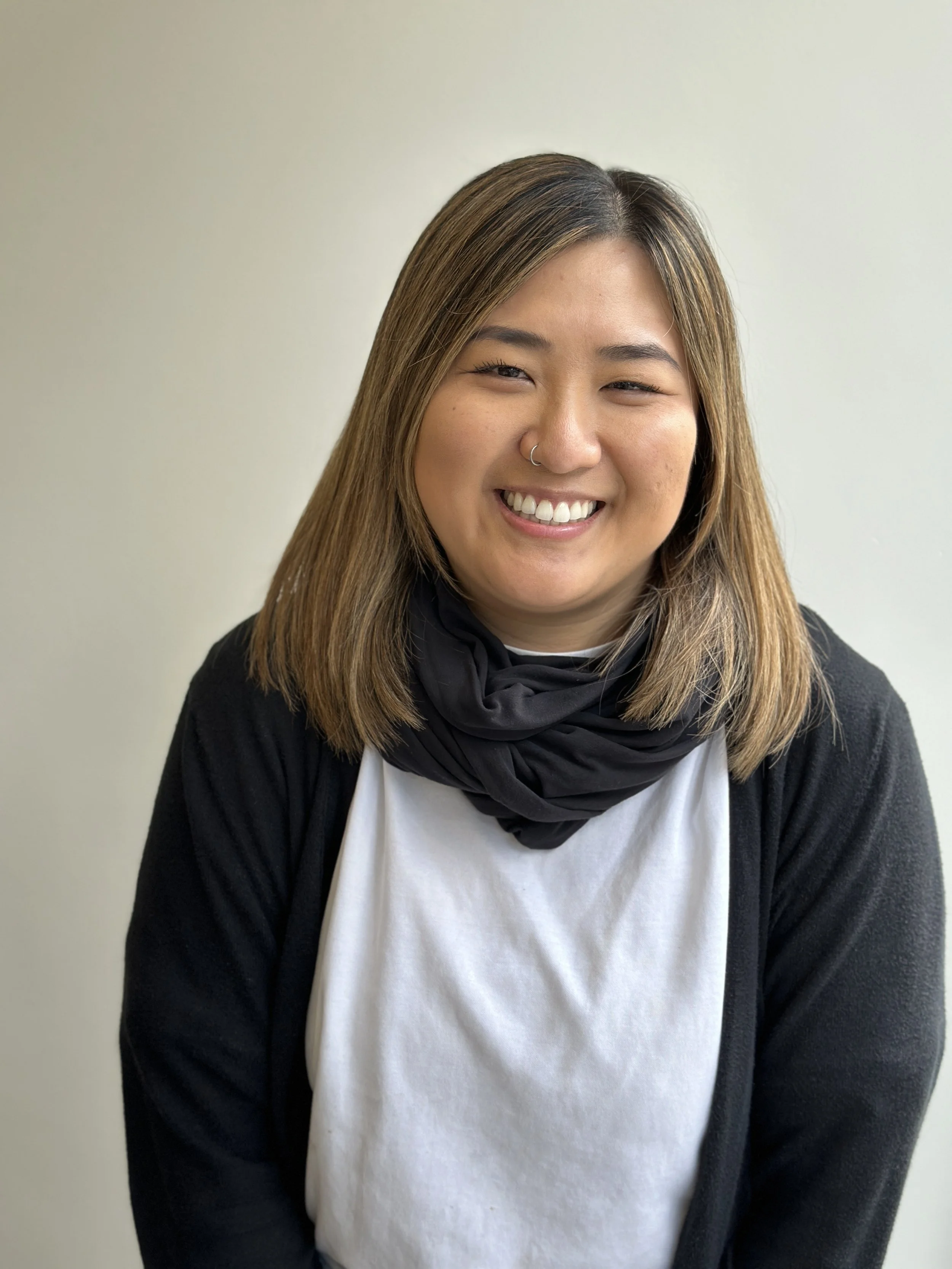A young woman with shoulder-length brown hair, smiling, wearing a black scarf, white shirt, and black cardigan, standing against a plain light-colored background.