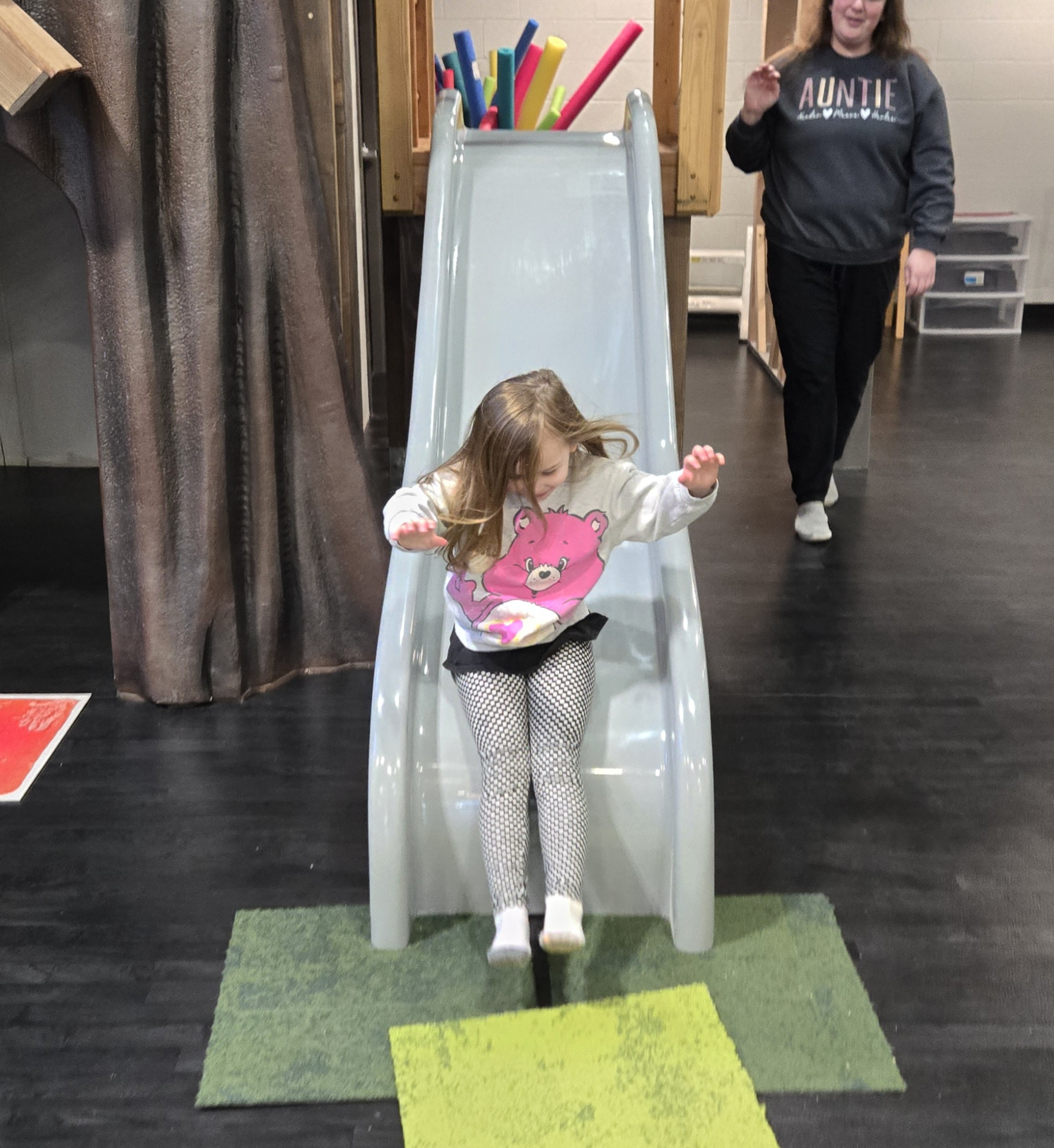 A young girl sliding down a small indoor slide with a white surface, on a patch of green and yellow carpet, while a woman in a gray sweatshirt watches nearby. The girl is wearing a white hoodie with a pink bear graphic and black and white checkered pants.