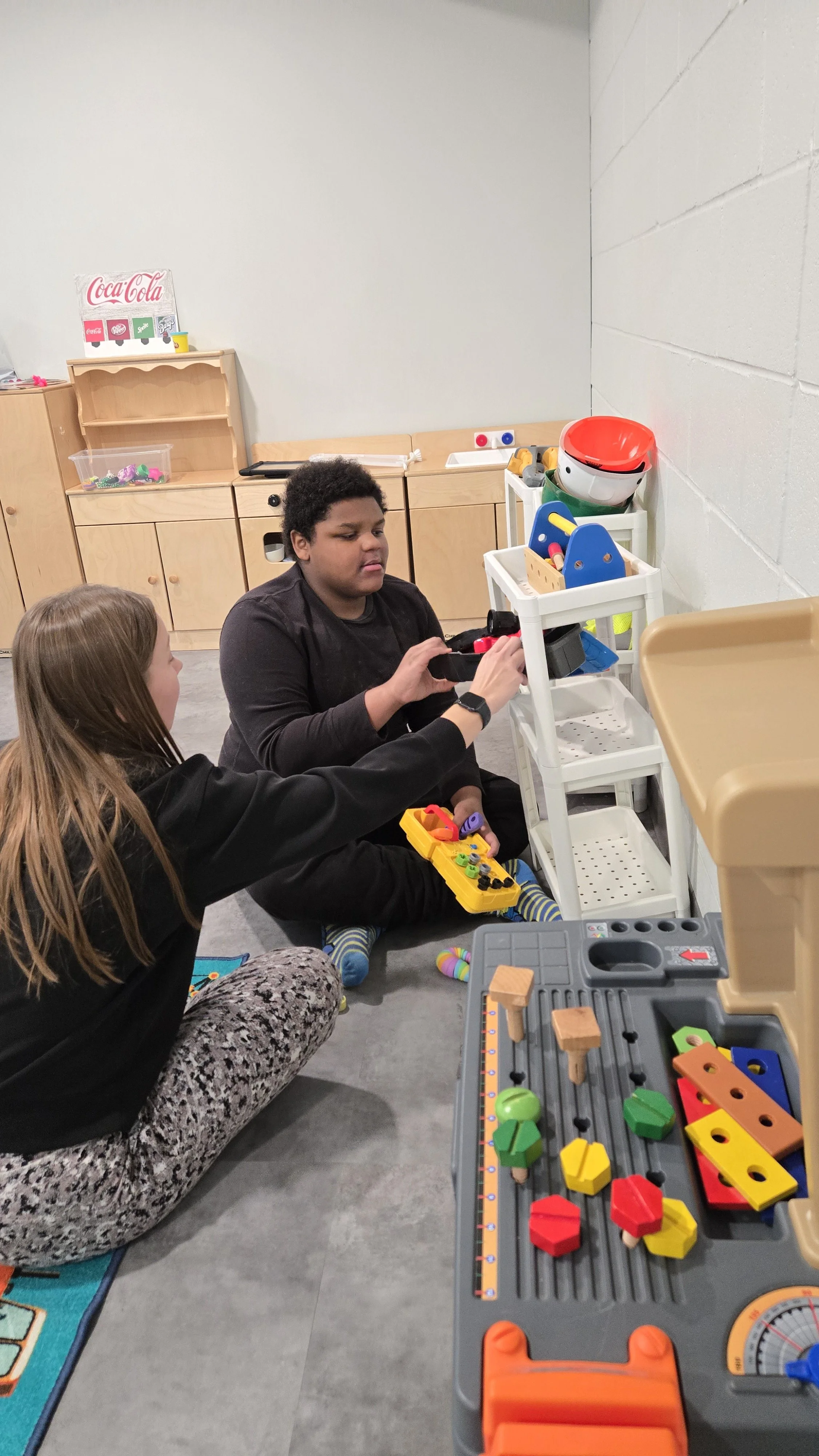 Two children playing with toys on the floor in a playroom, with shelves and a toy cart in the background.