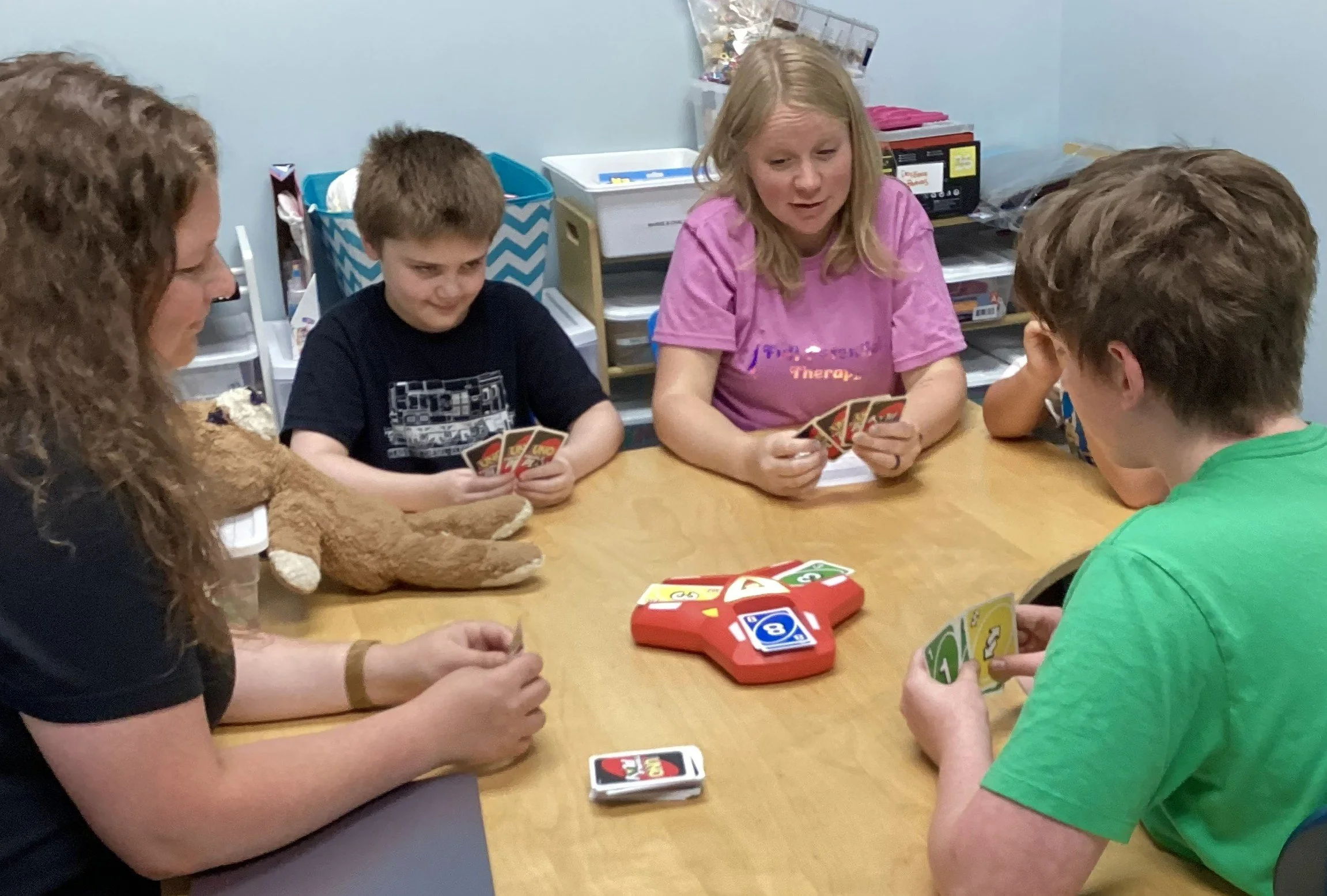 A group of children and a woman playing a card game at a wooden table in a classroom or activity room.