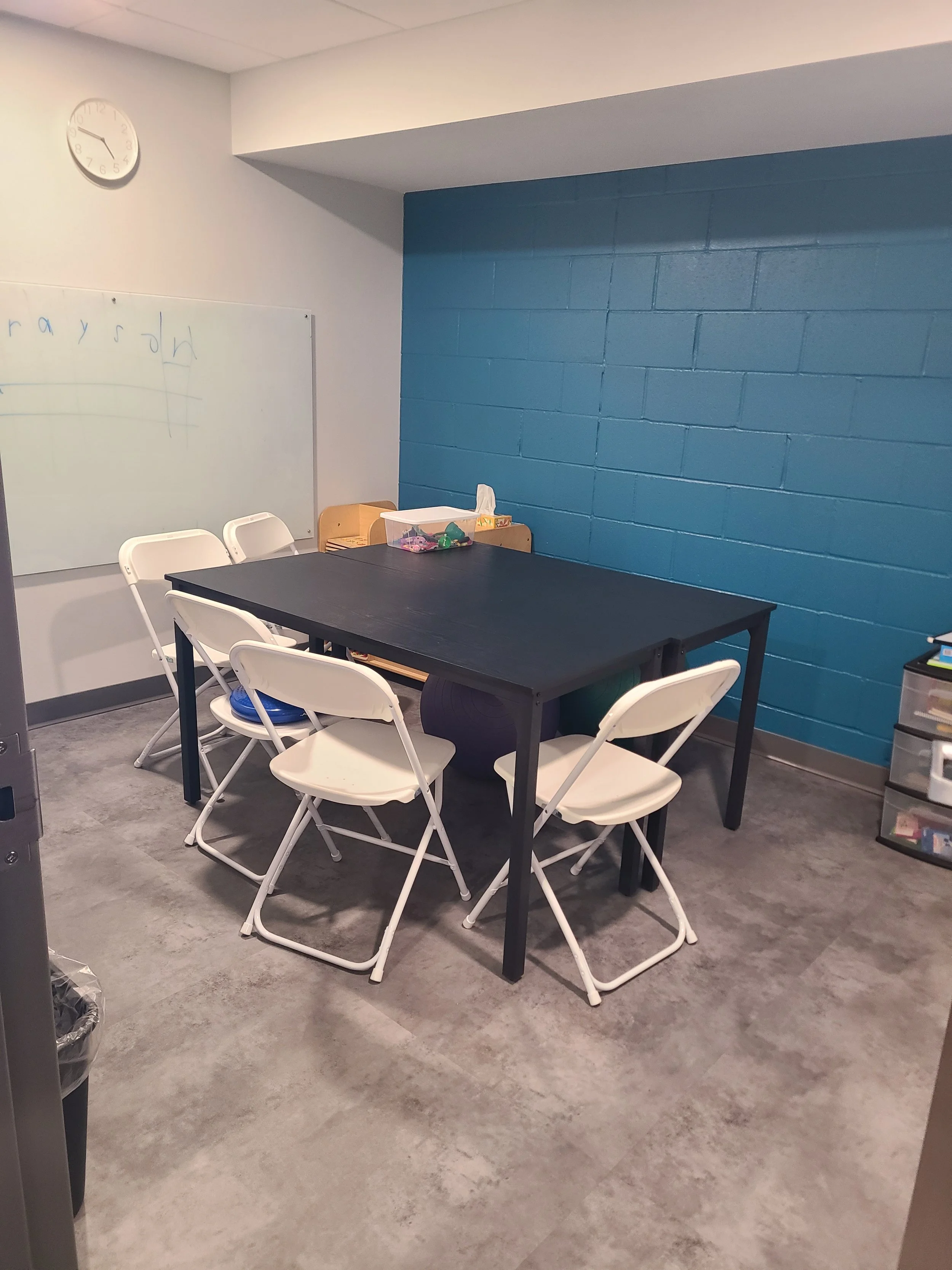 A small conference or meeting room with a black table surrounded by white folding chairs. A whiteboard with faint writing and a clock are on the wall, with a blue painted brick wall on one side. There are plastic storage bins and small supplies on a bench against the wall.