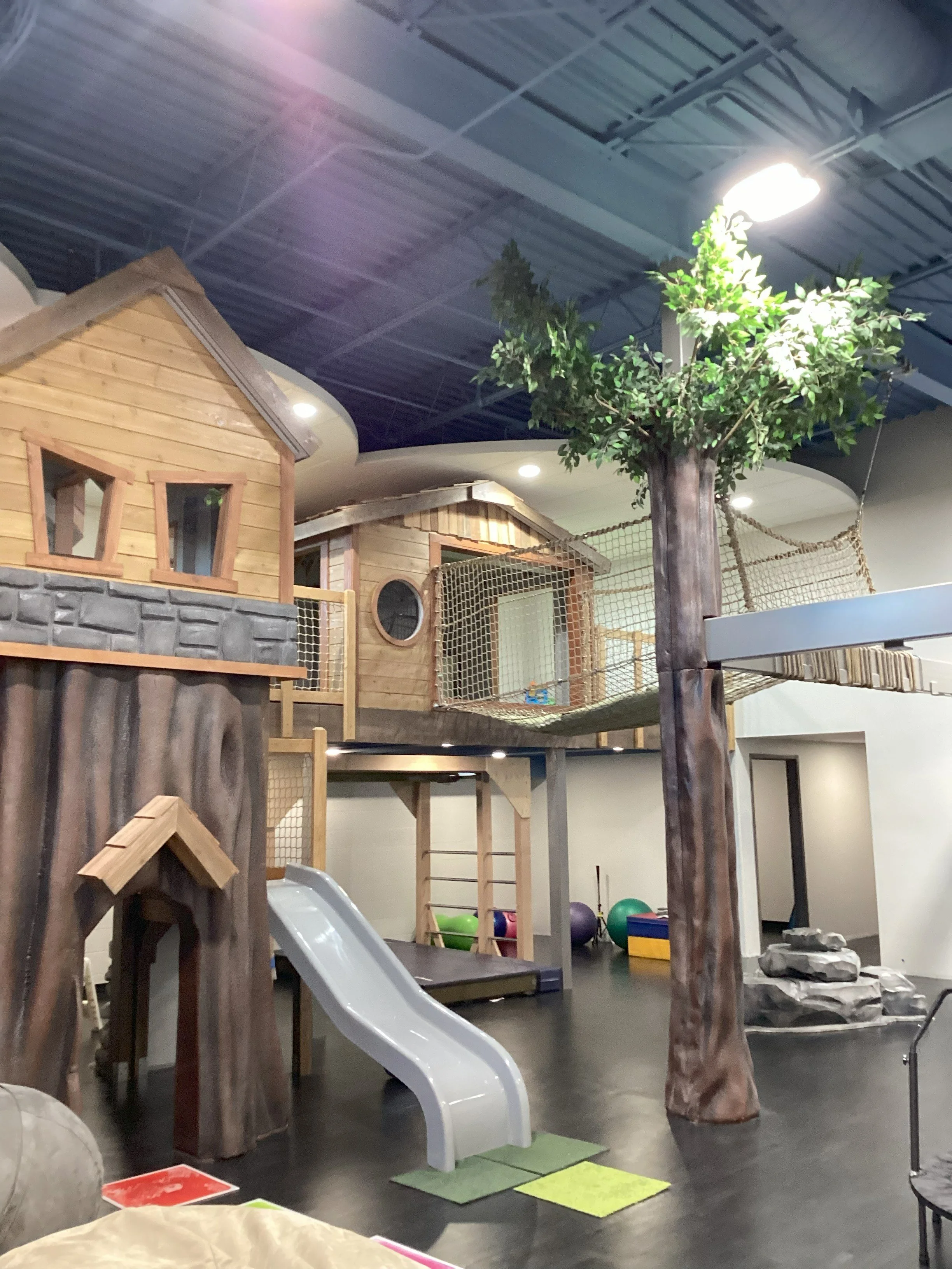 Indoor playground area featuring a wooden house structure with a slide, artificial trees, and various play equipment for children.