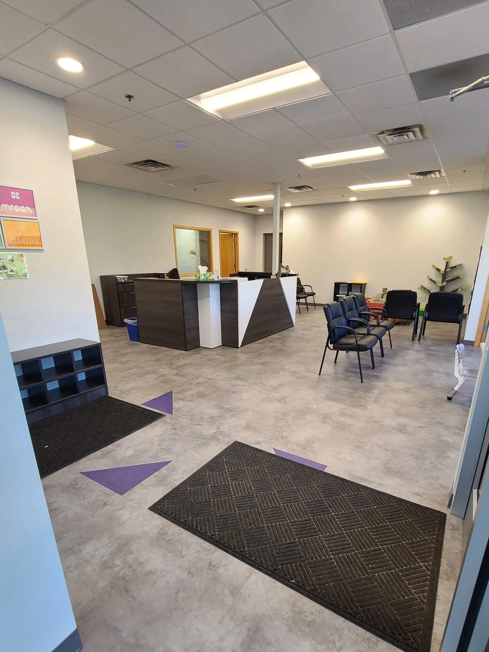 An office lobby with a reception desk, chairs, a plant, and decorative floor mats.