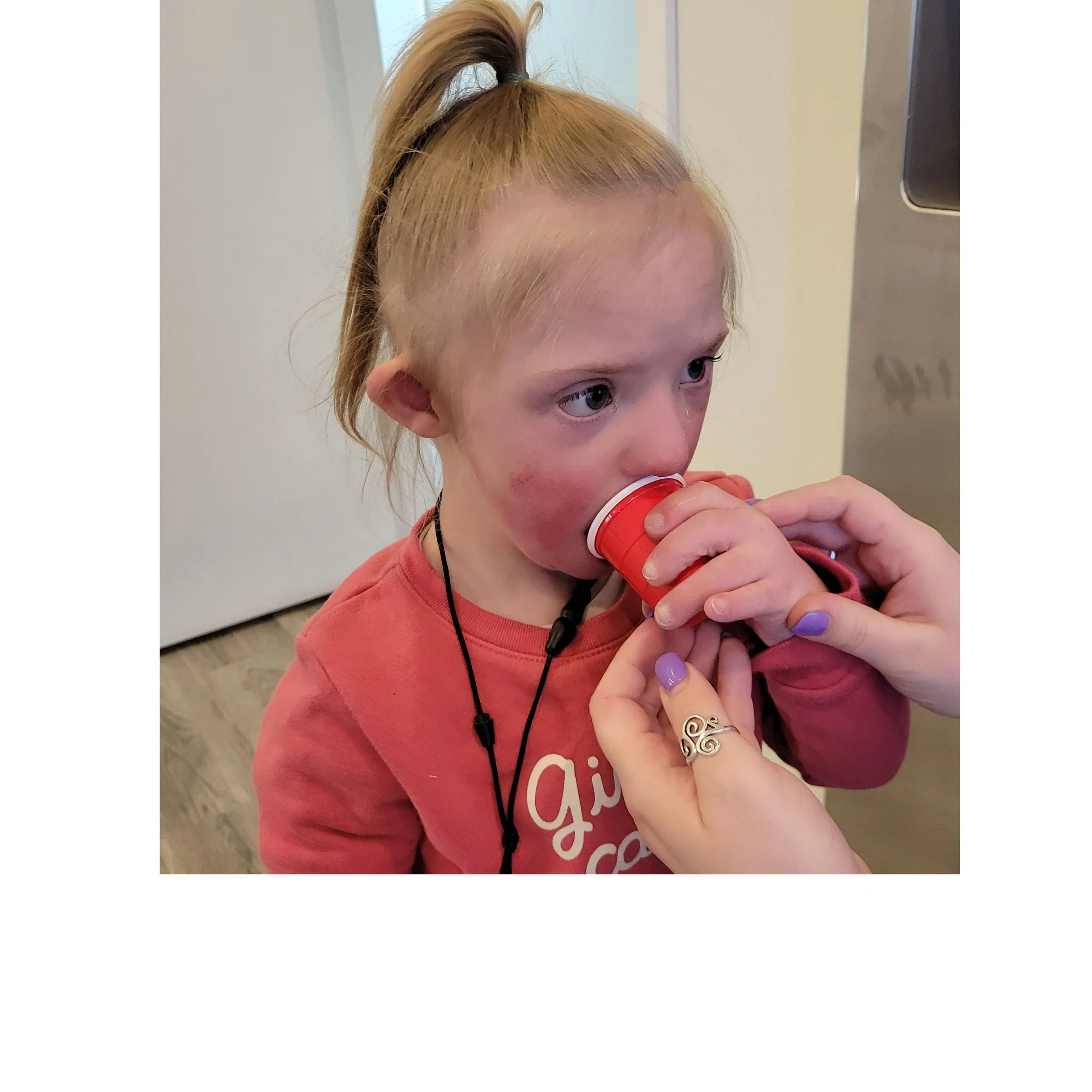 A young person with strawberry blonde hair tied up in a high ponytail, holding a red and white cup near their mouth, with purple painted nails, wearing a pink shirt with white writing, and a black necklace.