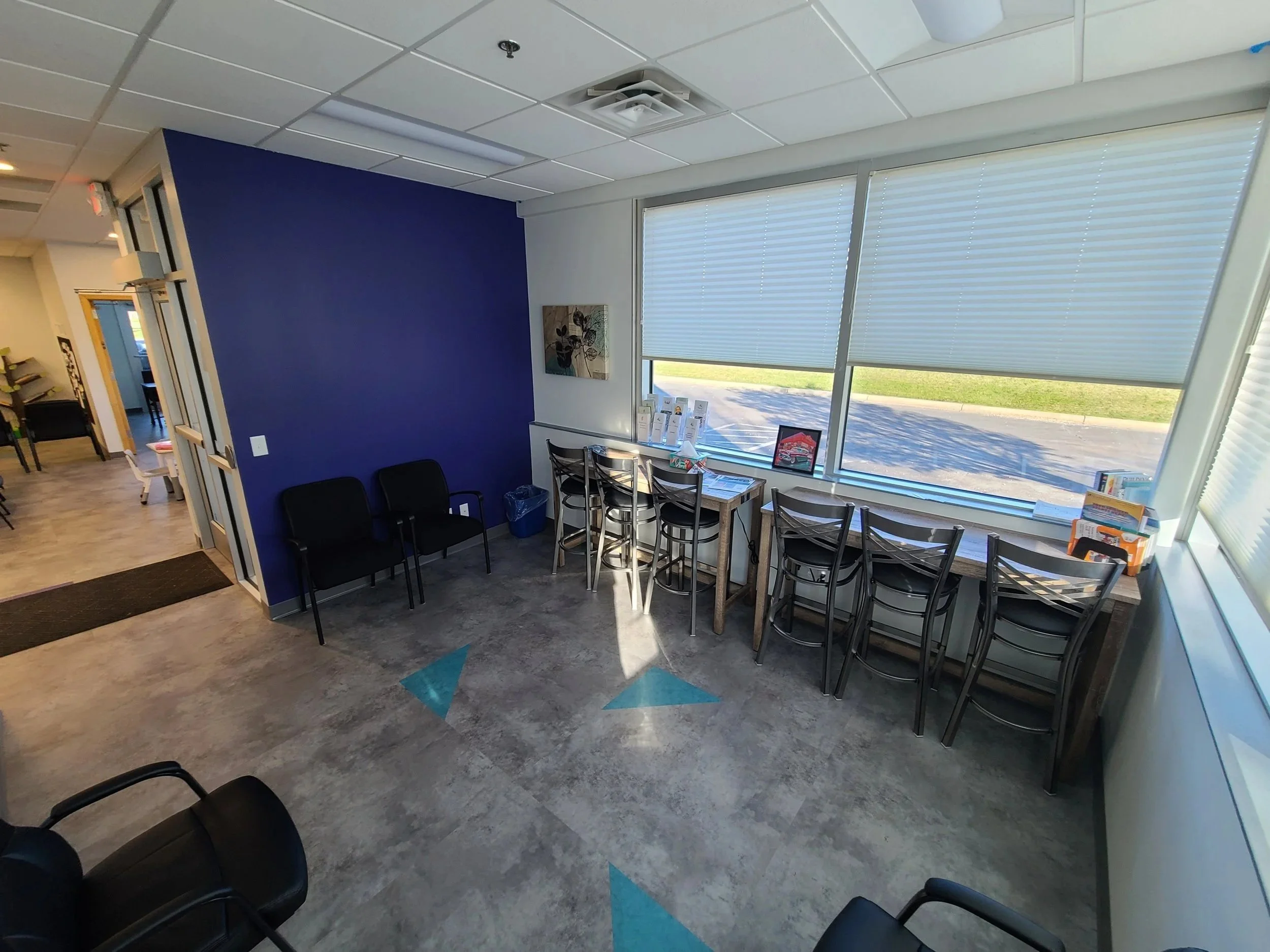 Interior of a waiting area with purple accent wall, black chairs, a small table with pamphlets, large windows with blinds, and a view of a grassy outdoor area.