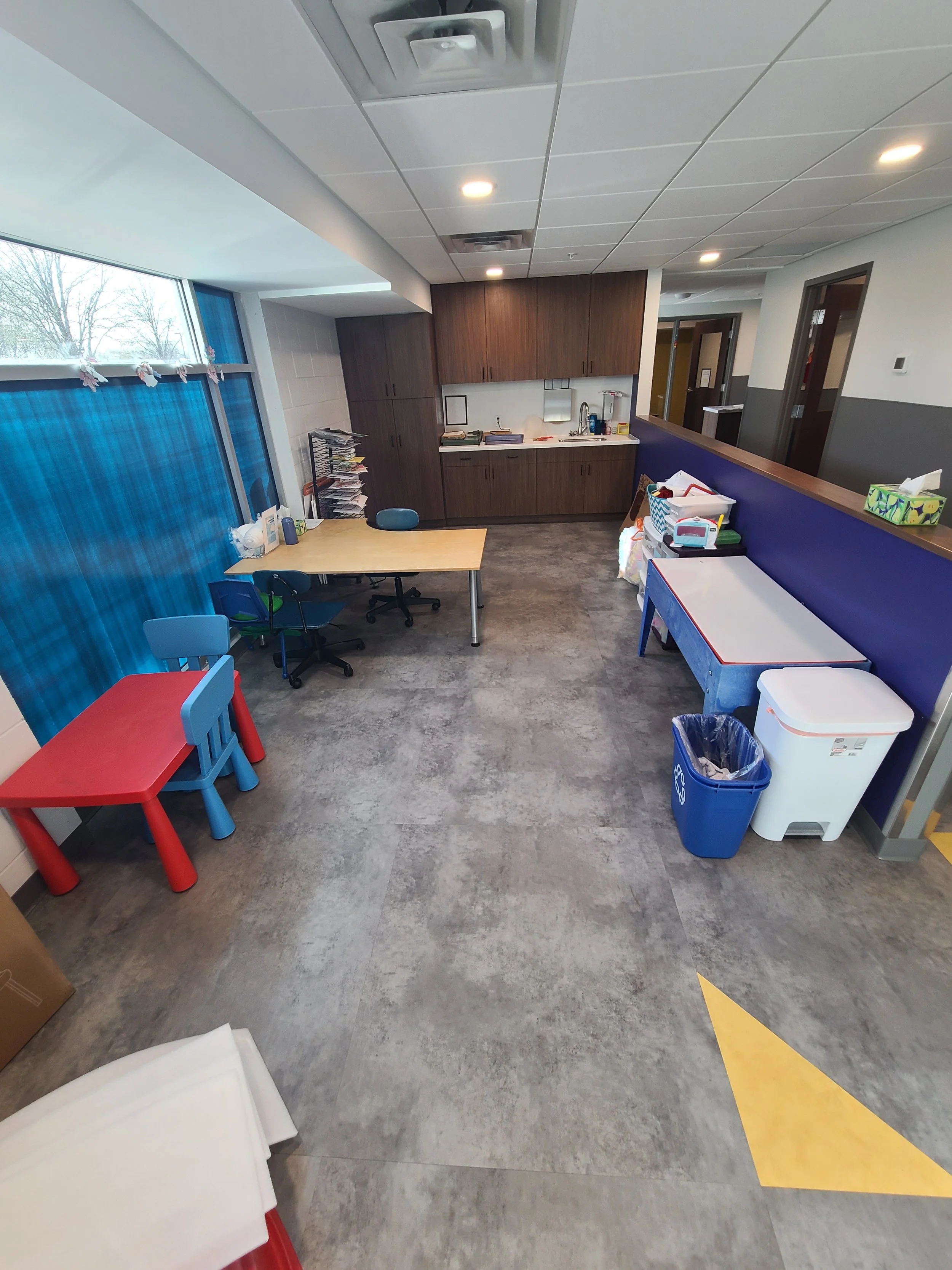 Empty medical or office break room with counter, cabinets, small table, chairs, waste baskets, and supplies.
