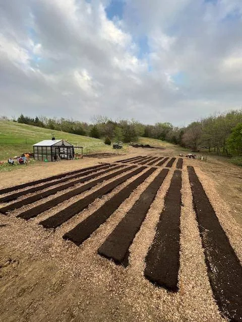 Garden beds with freshly tilled dark soil in a rural landscape, with a greenhouse and trees in the background under a partly cloudy sky.
