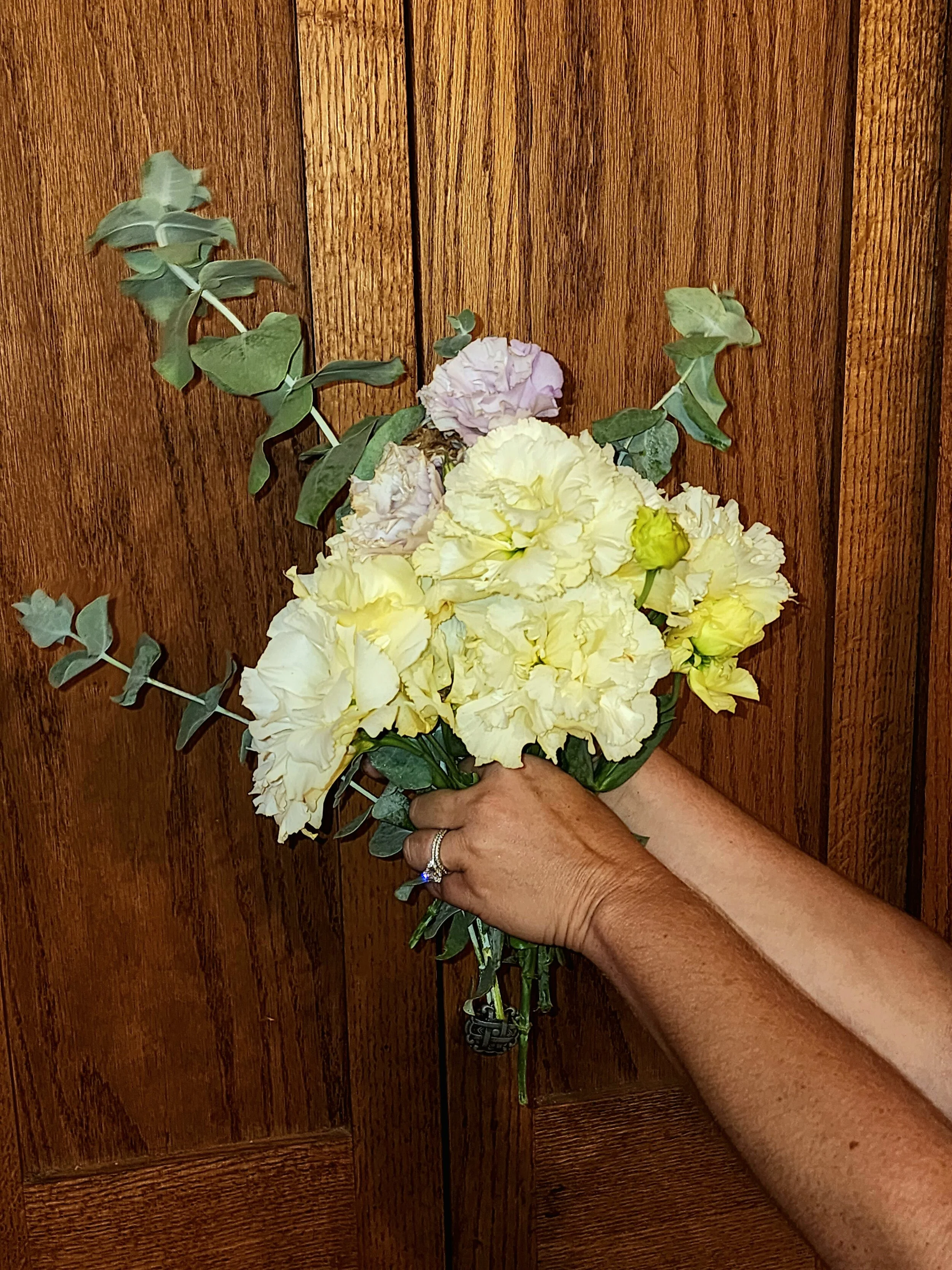 Person holding a bouquet of cream and lavender flowers with green leaves in front of a wooden background.