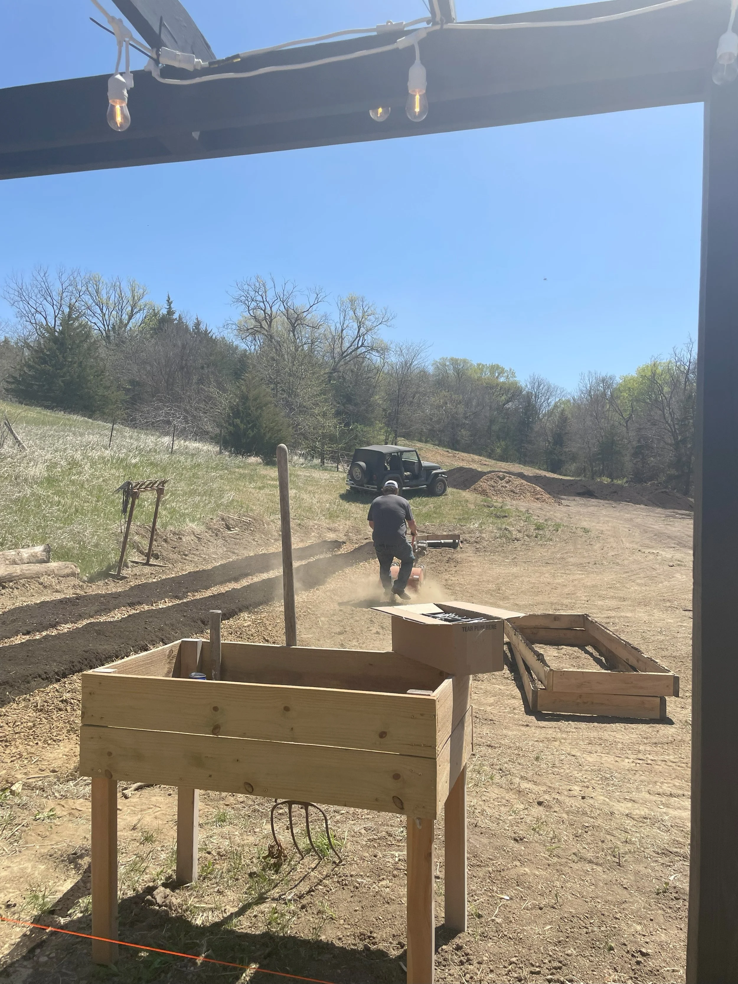 Man working outdoors on a sunny day, digging or working with a lawn roller on a dirt path near trees, a vintage black vehicle, and wooden structures in the foreground.