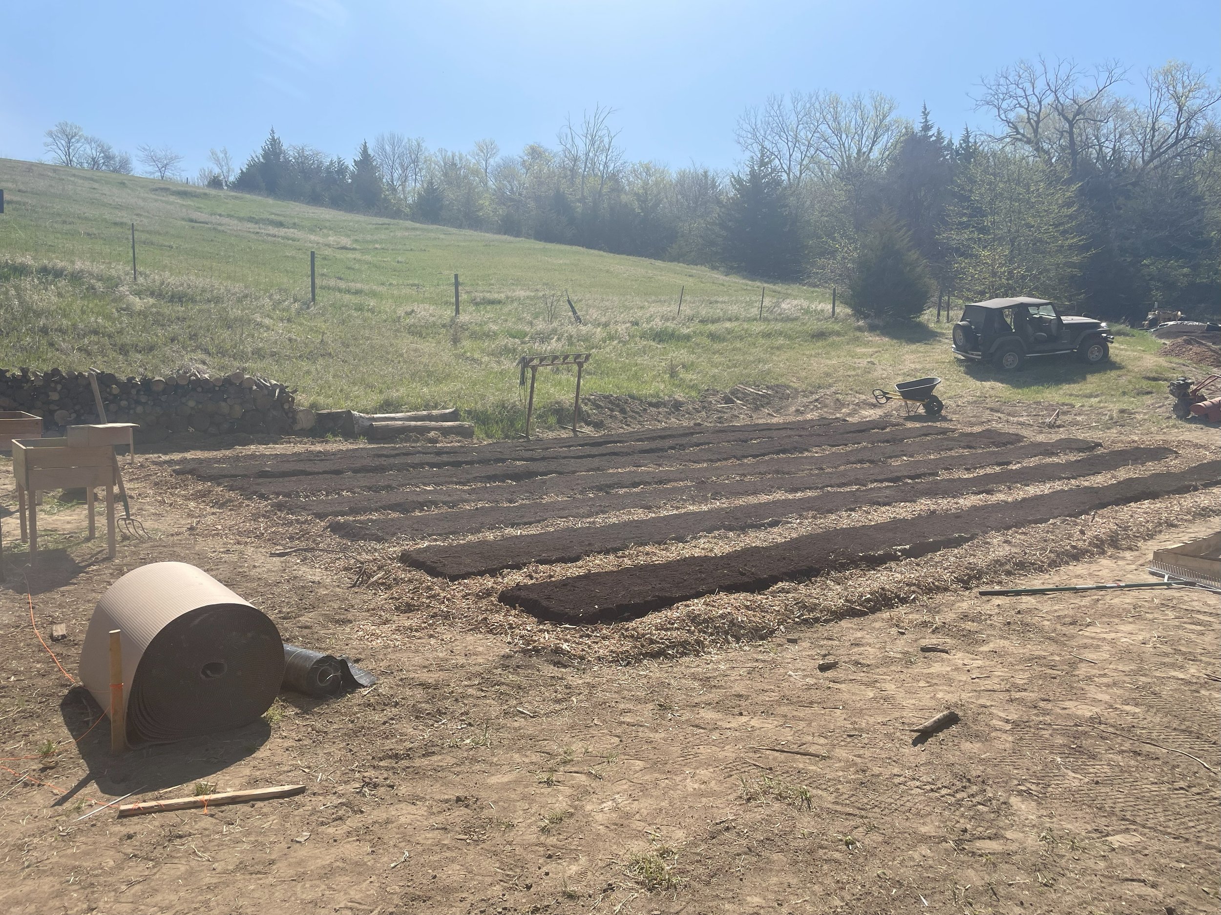 Freshly tilled garden beds on a farm with trees and a hill in the background, a tractor, wheelbarrow, and gardening tools visible.