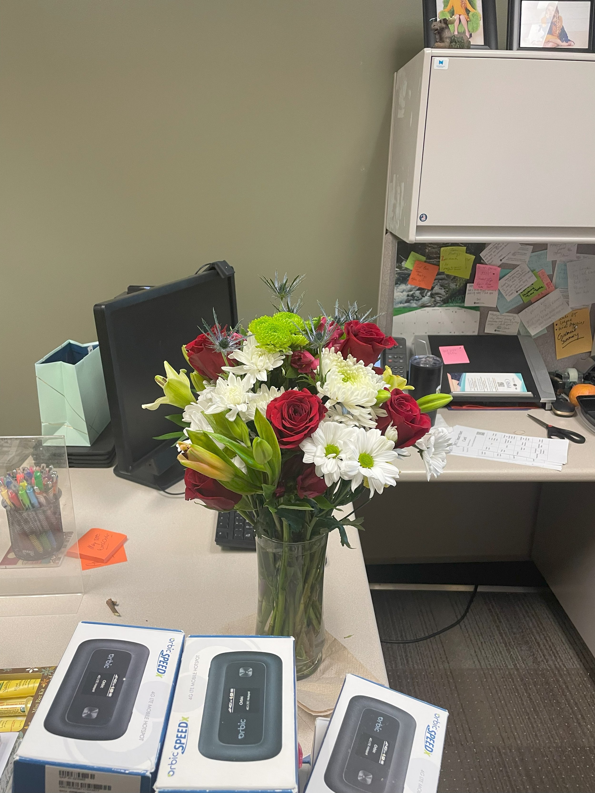 A desk with a bouquet of red roses, white daisies, and green flowers in a glass vase in the center. Surrounding the bouquet are office items including boxed portable Wi-Fi devices labeled 'Optic,' a computer monitor, sticky notes, pens, papers, and p