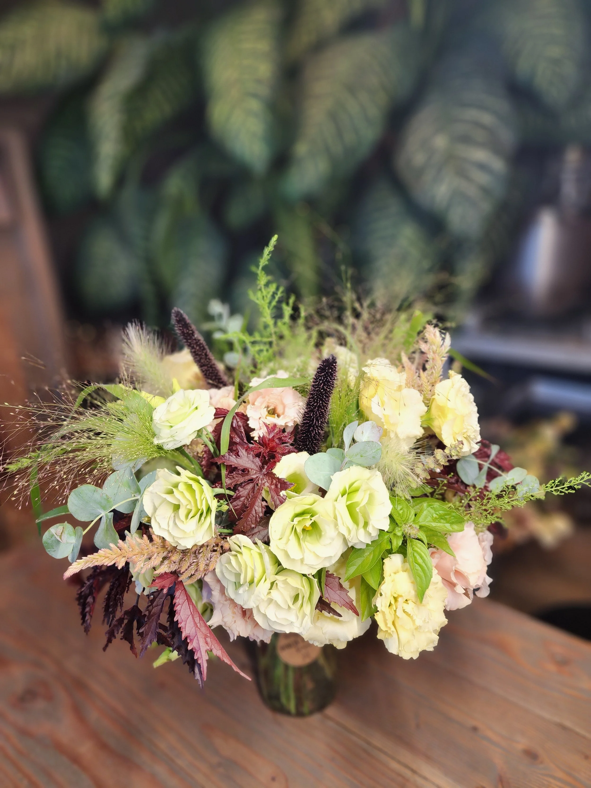 A floral bouquet in a glass vase featuring white, pale yellow, and light pink flowers along with greenery and textured dark purple accents, set on a wooden surface with a blurred background of patterned green leaves.