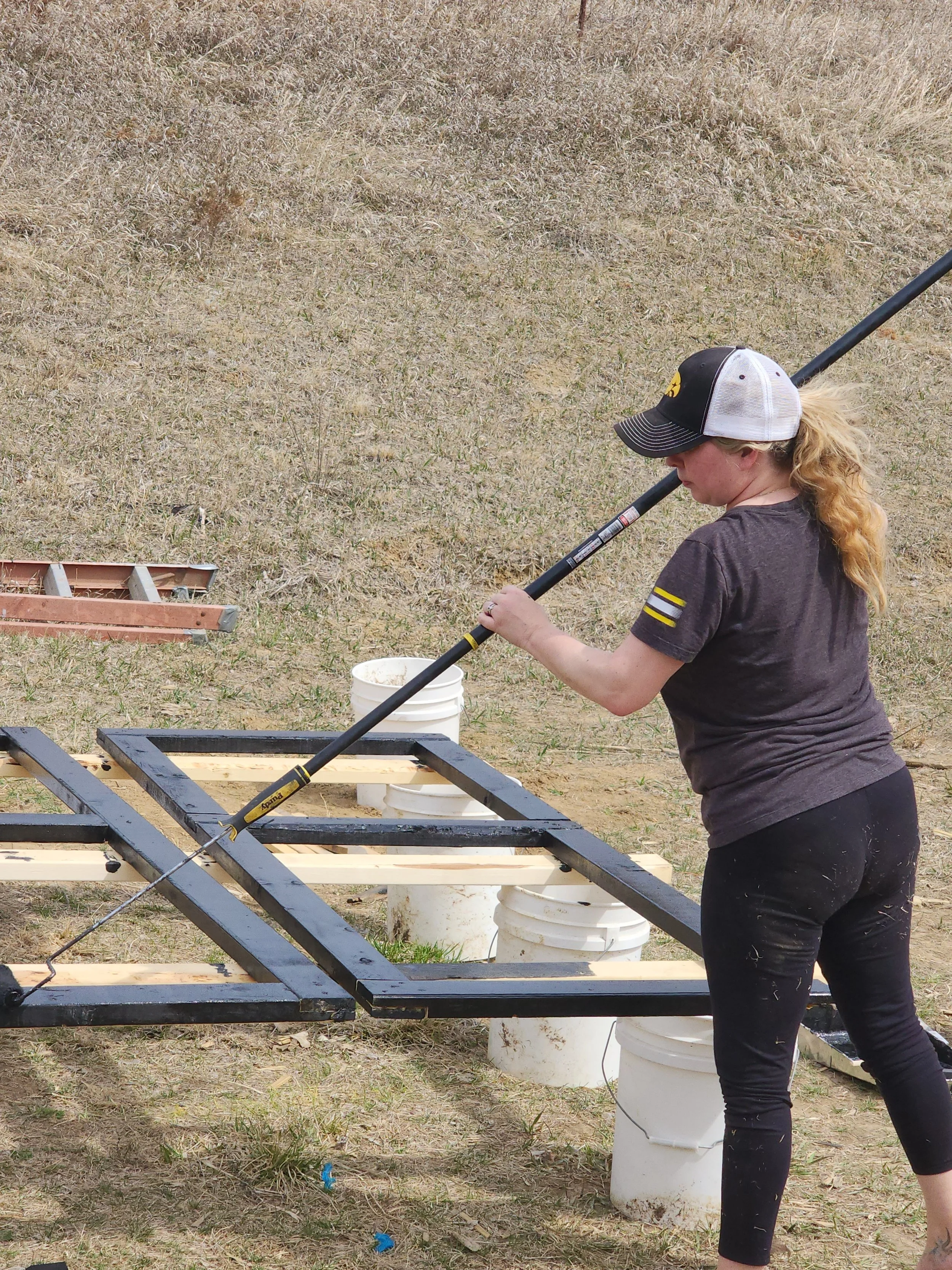 A woman working outdoors, using a long tool on a black metal frame supported by white buckets.