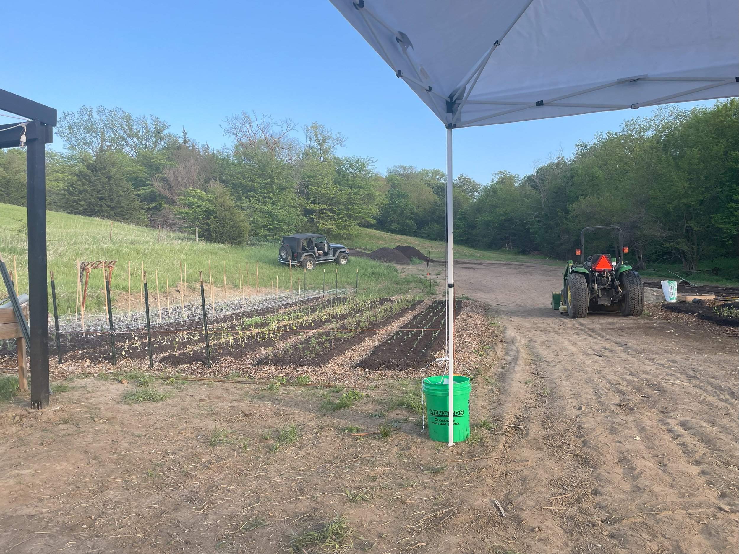 A small farm or garden with planted rows of seedlings, a tractor, and a pickup truck on a dirt road against a background of green trees, under a partly cloudy sky, seen from beneath a white canopy.