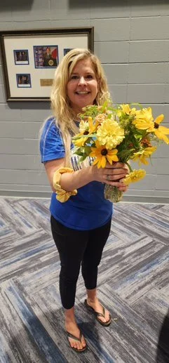A woman with blonde hair smiling and holding a bouquet of yellow flowers in an indoor setting, with framed pictures on the wall behind her.