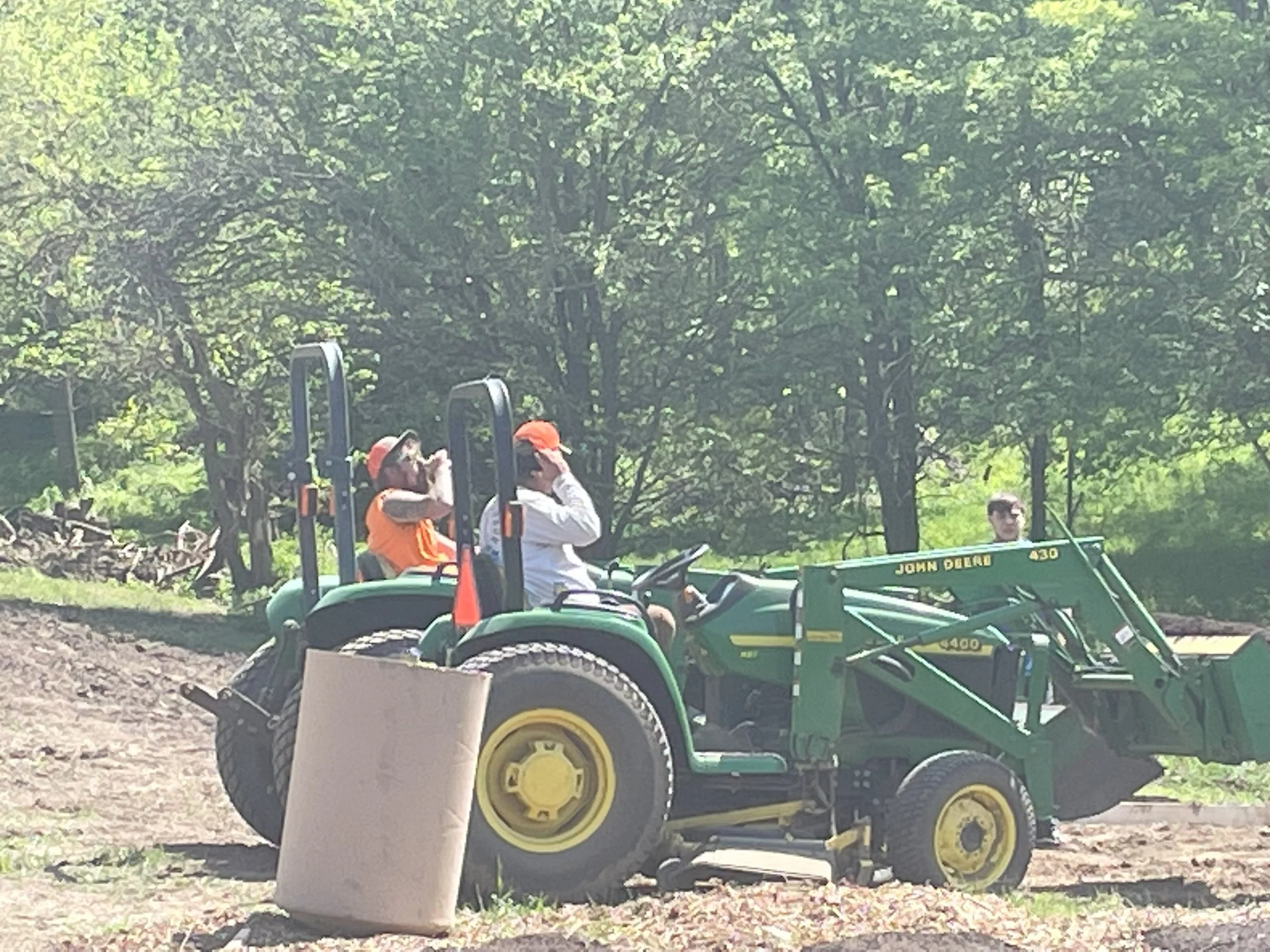Two people sitting on a John Deere tractor, one wearing an orange shirt and hat, the other in white with an orange cap, in a green outdoor area with trees.