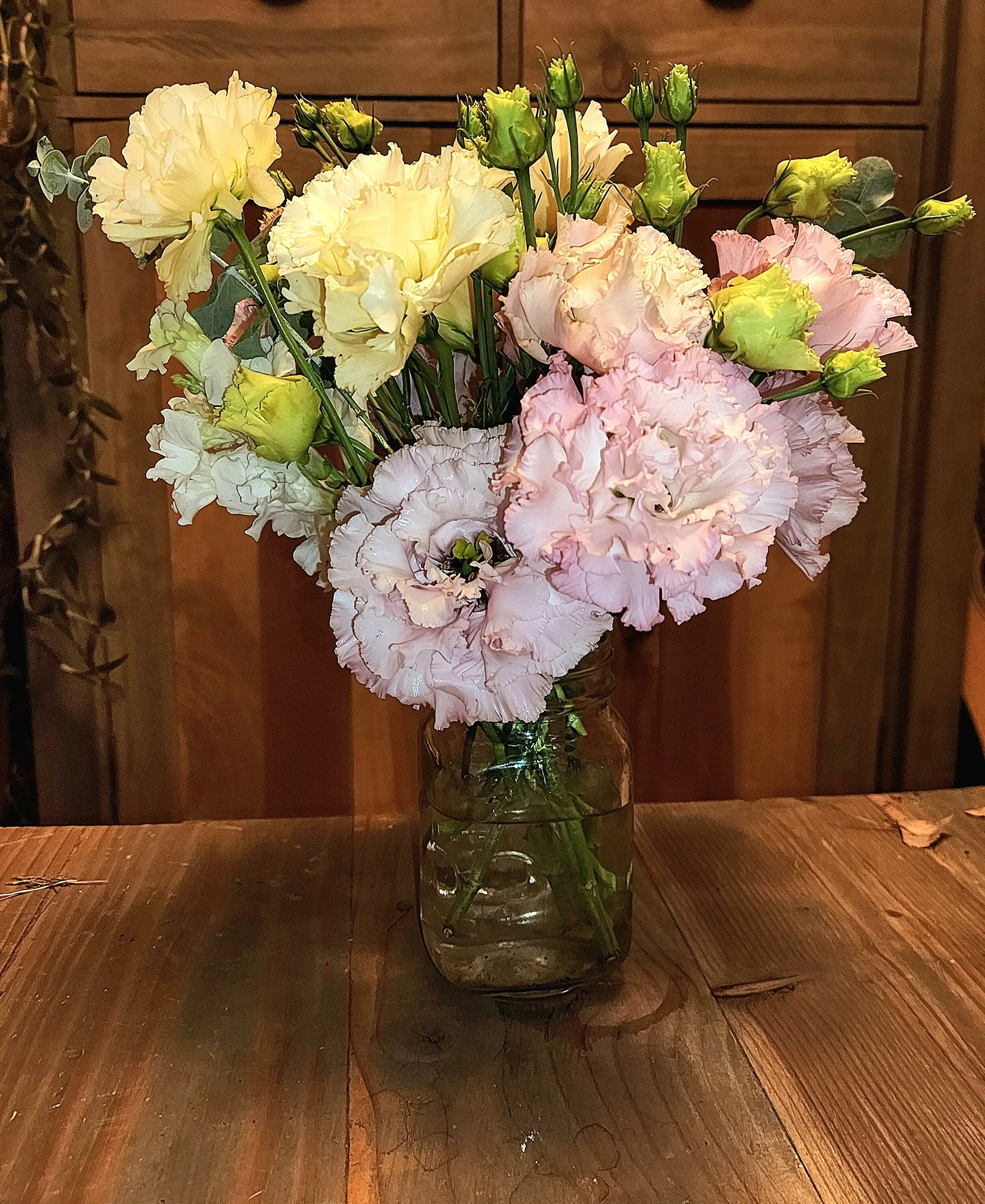 A glass jar filled with water holding a bouquet of light pink, white, and pale yellow flowers, set on a wooden table with a wooden cabinet in the background.