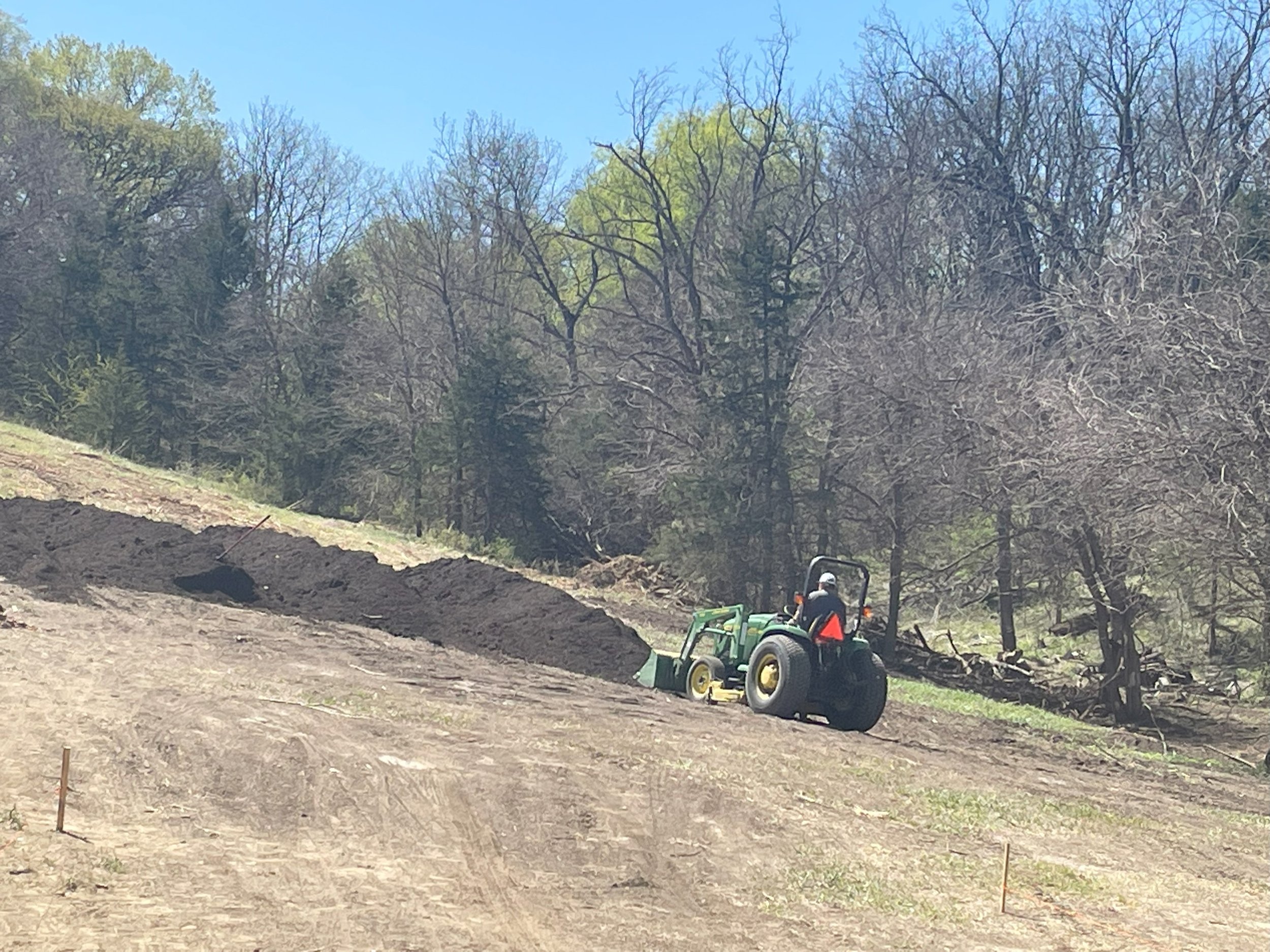 A person operating a green tractor on a dirt hillside, surrounded by leafless trees in a rural outdoor setting.