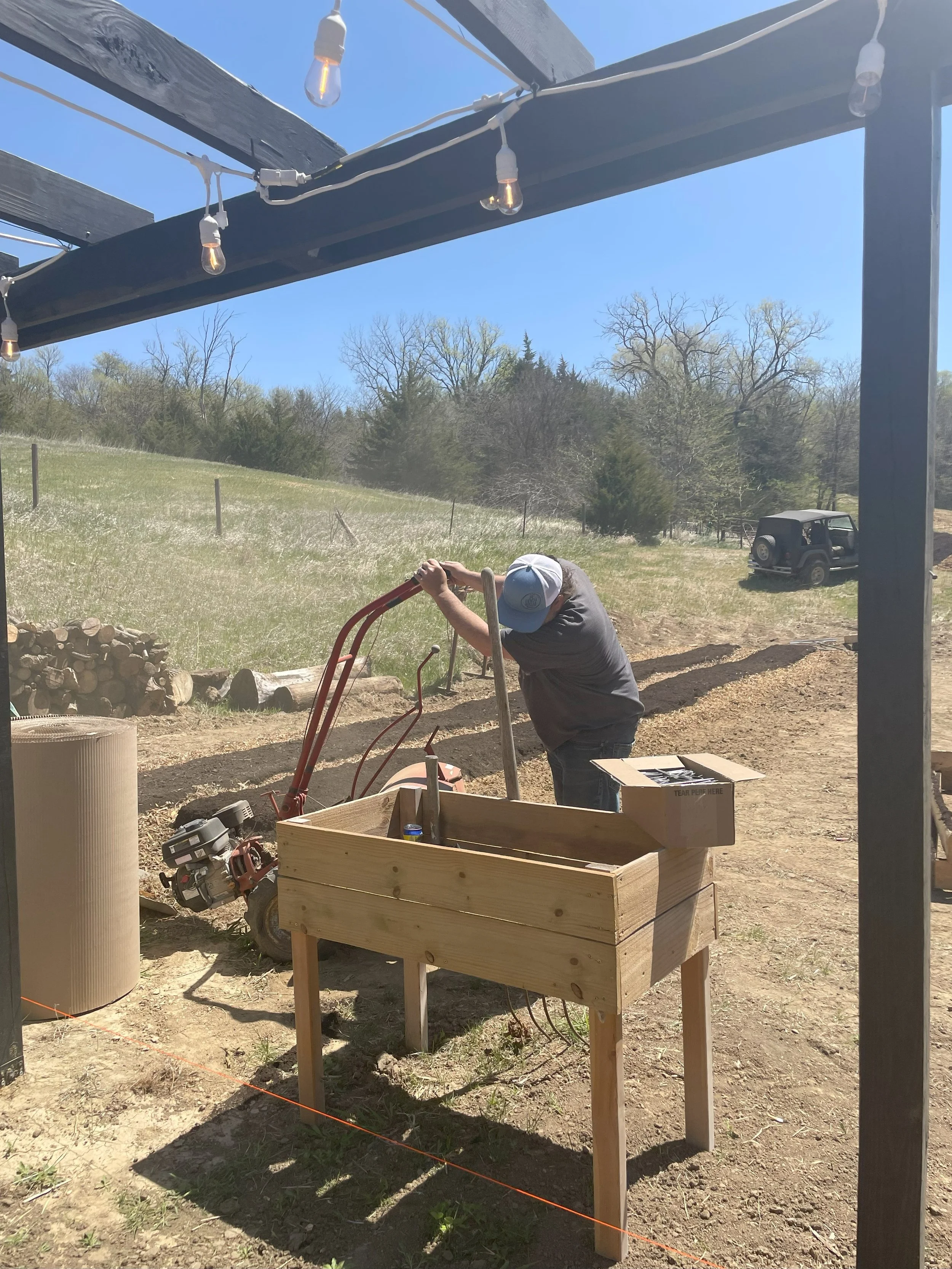 A person wearing a blue and white cap is using a manually operated tiller or rototiller in a field. There is a wooden table in the foreground, a box on it, and an open outdoor landscape with a clear blue sky and a jeep parked in the distance.