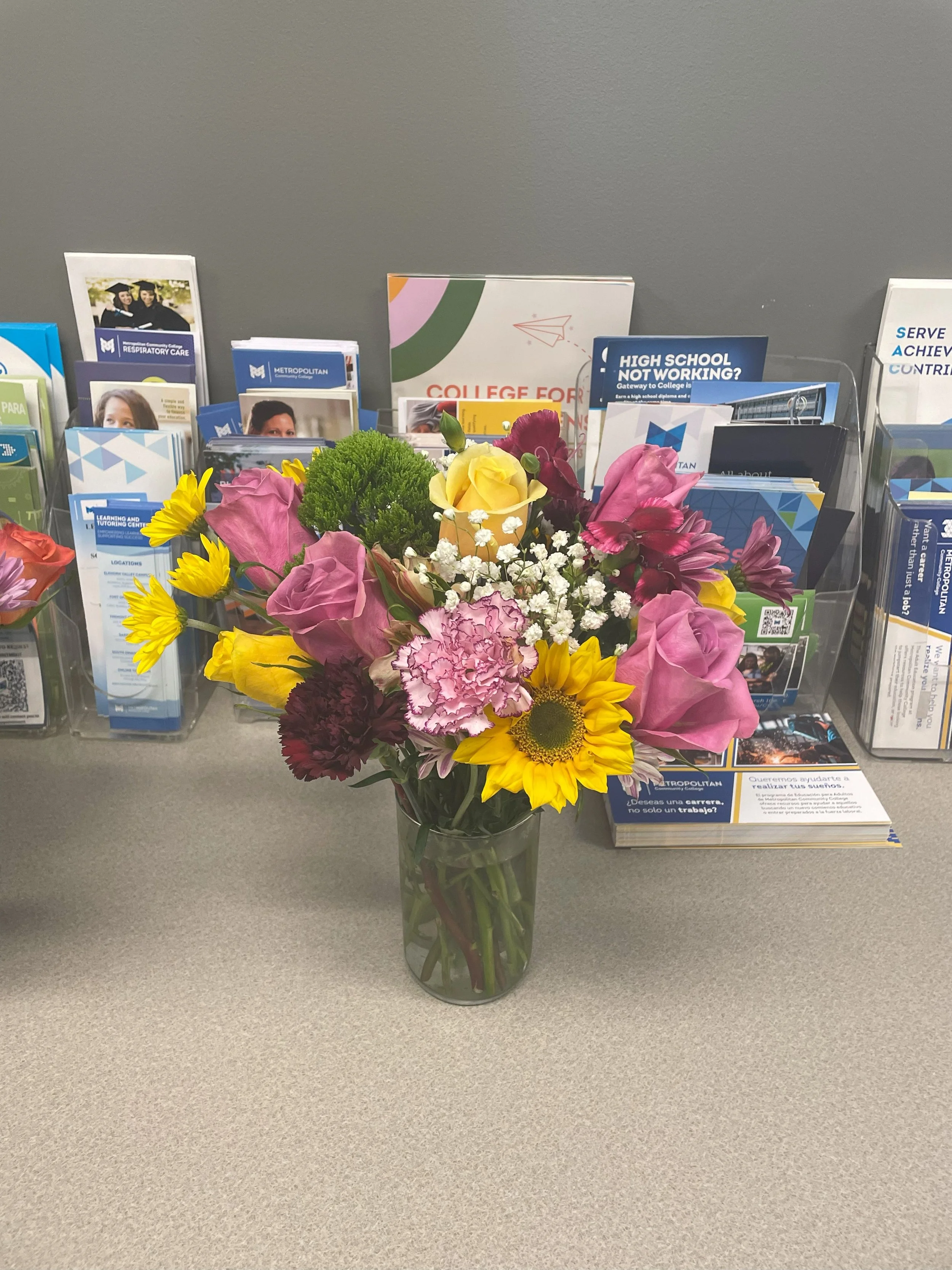 Colorful bouquet of flowers in a glass vase on a gray table in front of brochures and informational materials.