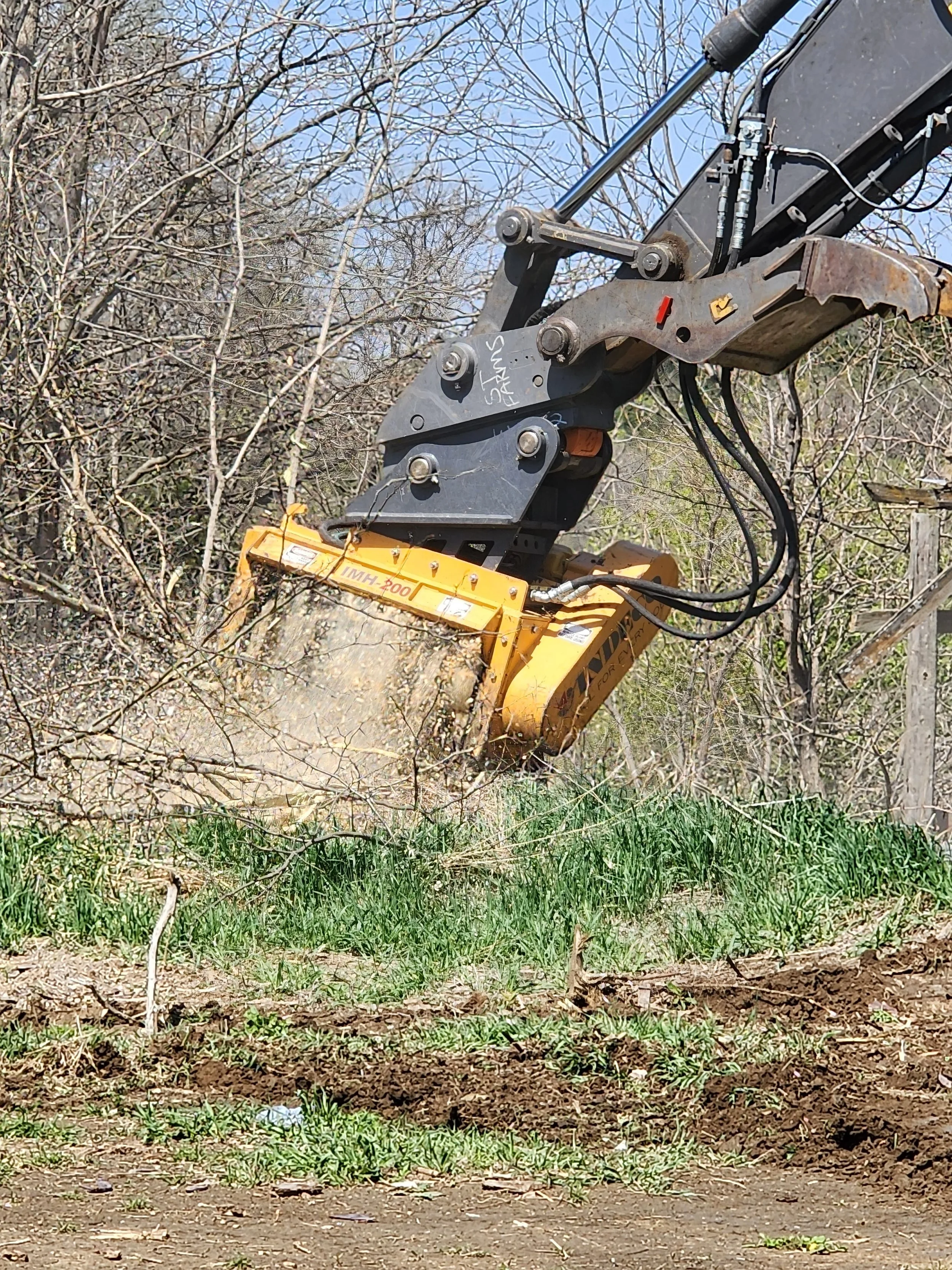 Close-up of a large yellow hydraulic excavator attachment breaking ground and digging into the dirt, with surrounding trees and blue sky in the background.