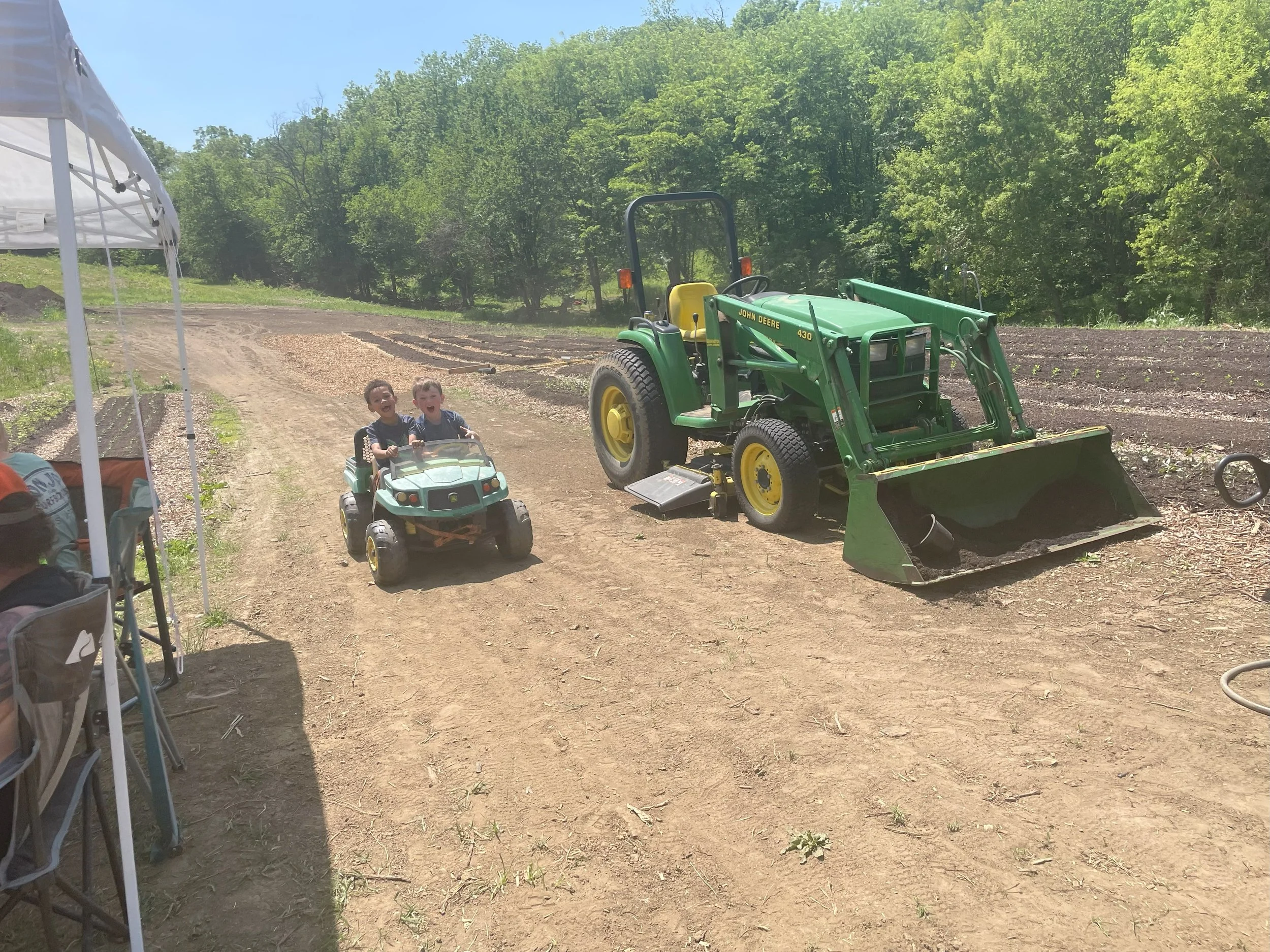 Two young boys riding a small toy car on a dirt path, with a green John Deere tractor parked nearby, in a rural outdoor setting with trees and cultivated fields in the background.