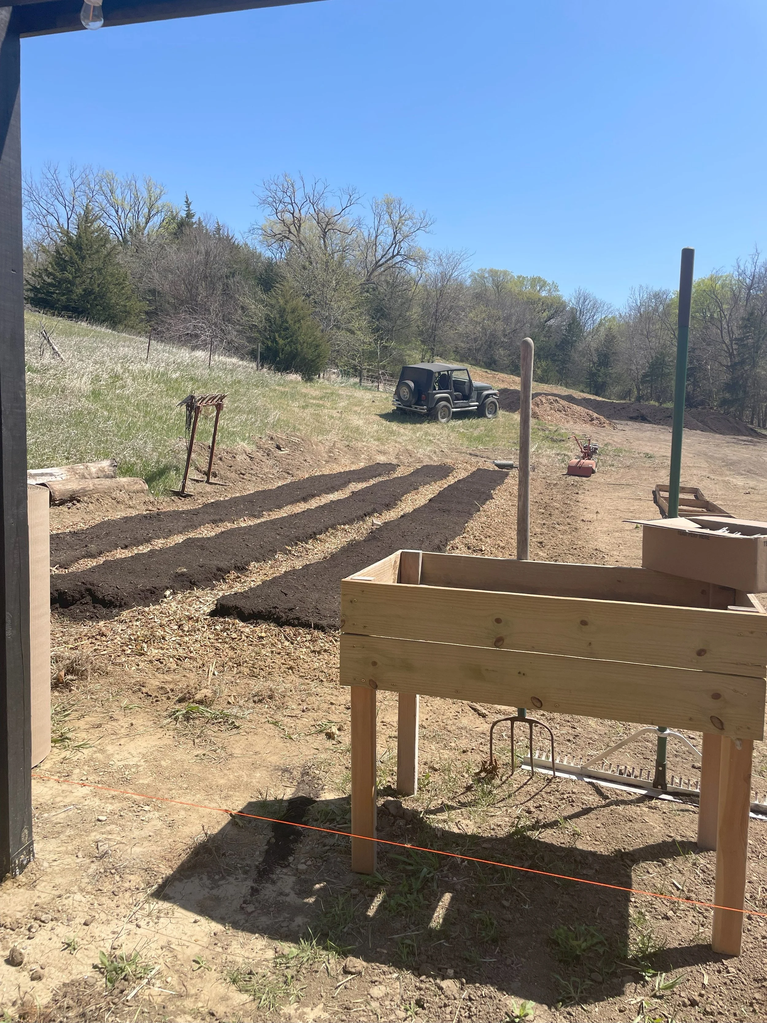 A garden plot with freshly tilled soil and four planting rows, set in an open yard with trees and a blue sky in the background. There is a black off-road vehicle parked on a hill in the distance. A wooden raised garden bed is in the foreground.