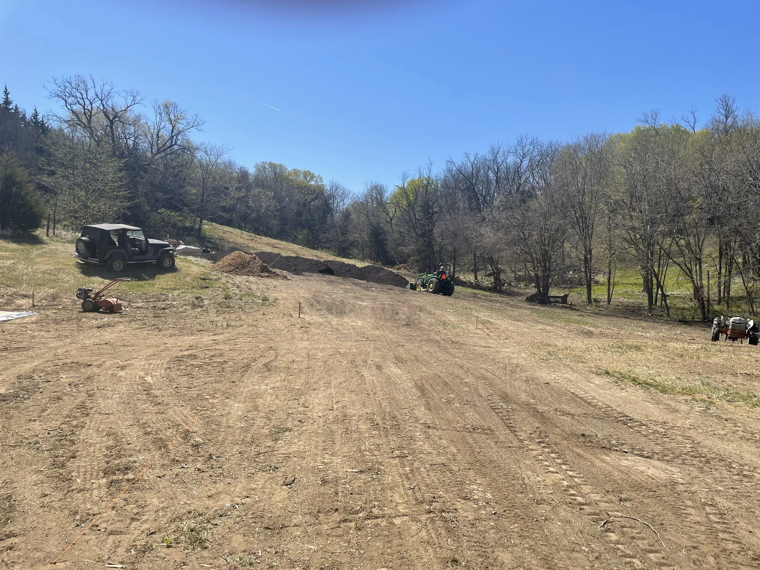 A cleared dirt area with construction equipment including a black Jeep, a lawnmower, and two tractors, surrounded by trees and a clear blue sky.