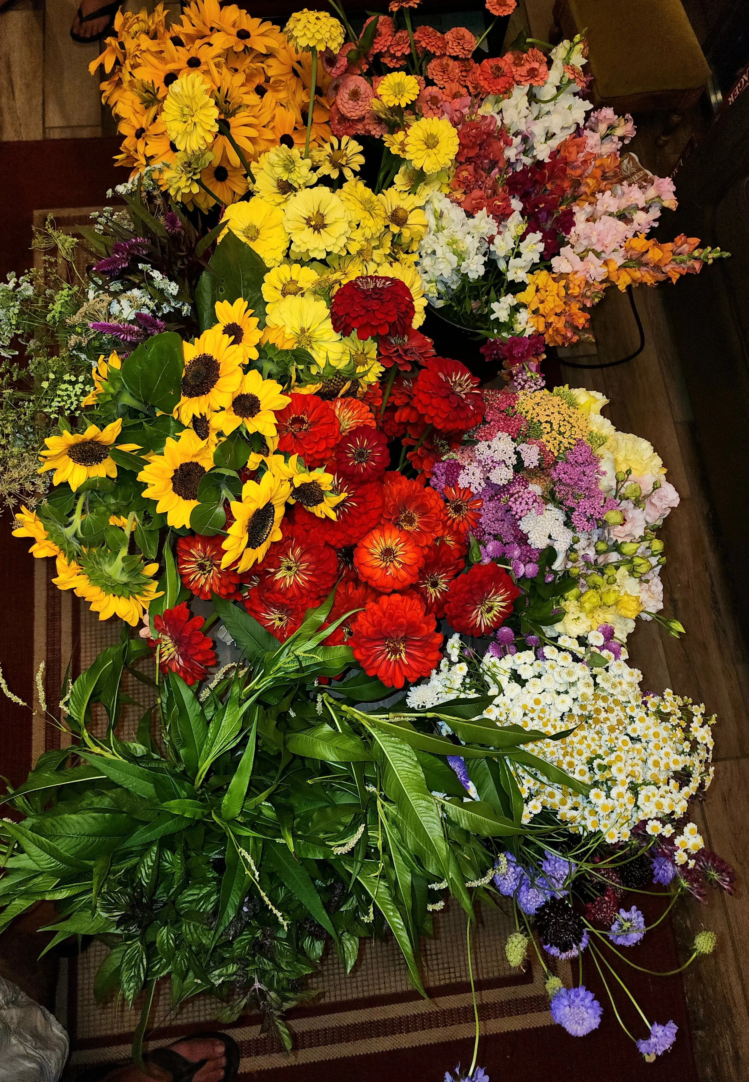 A variety of colorful flowers, including sunflowers, zinnias, daisies, and sweet peas, arranged in bouquets on a wooden table.